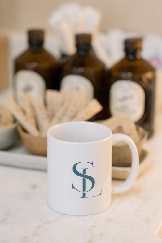 White ceramic mug with initials 'GSL' in blue, placed on a white surface. In the background, there are three brown bottles with black caps and a basket of paper napkins.