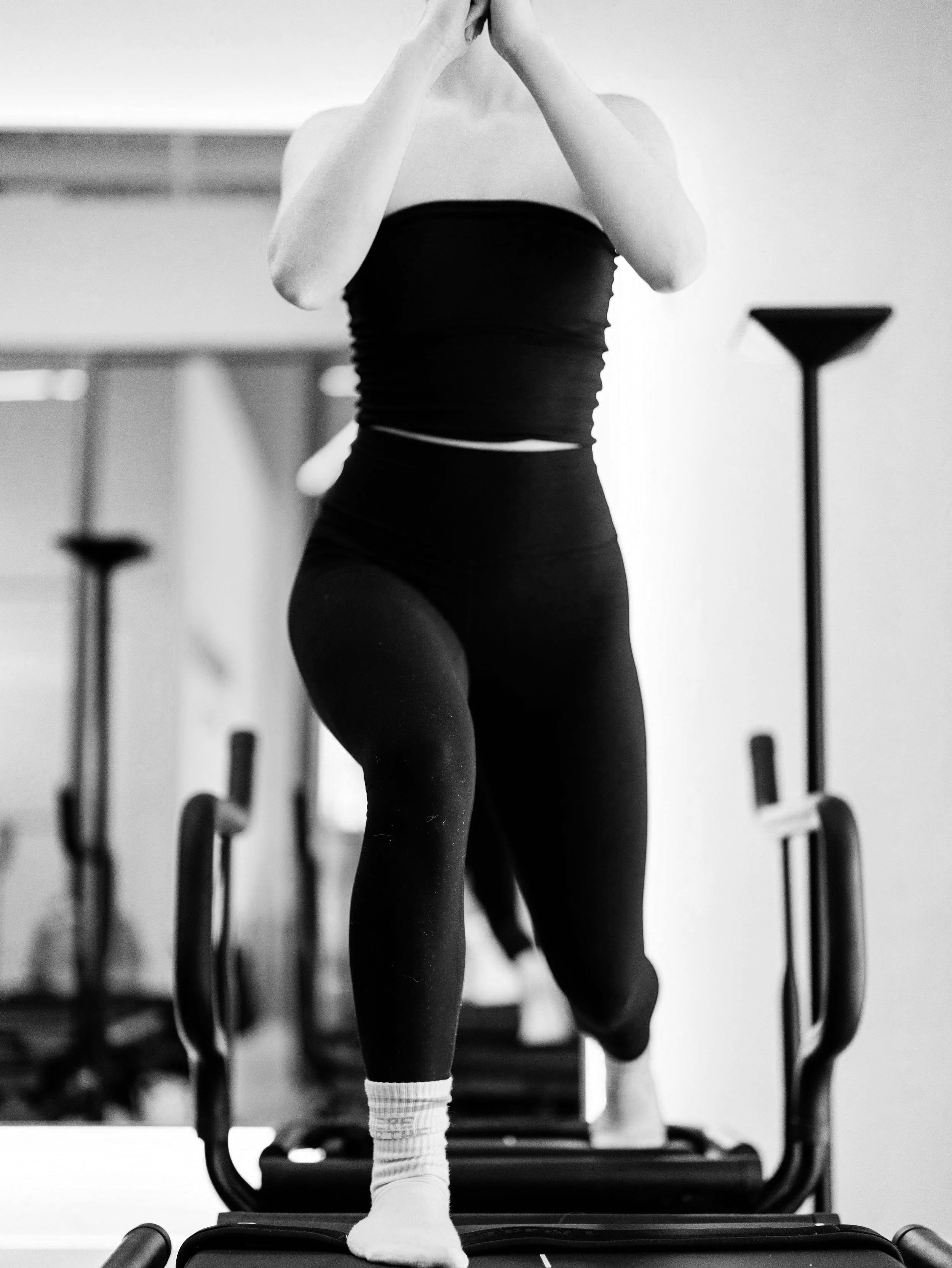 A woman lunging on a lagree mega reformer a gym, wearing a black sports bra, black leggings, and white socks.