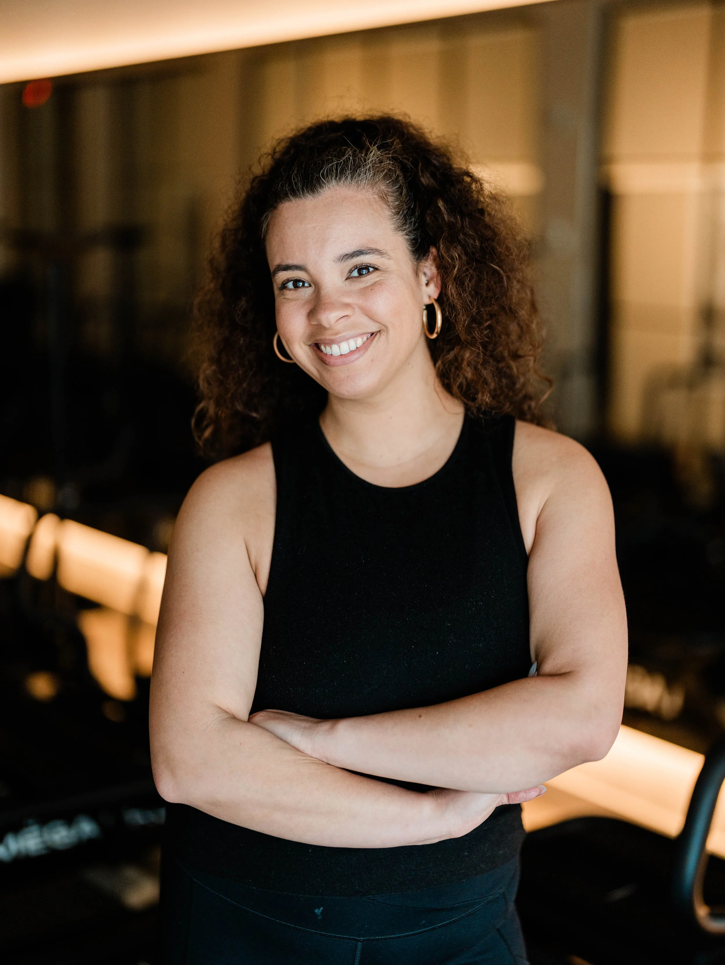 Raemi is a VersaClimber instructor with curly haid, wearing a sleeveless black top and gold hoop earrings, smiling with arms crossed in an indoor setting with warm lighting.