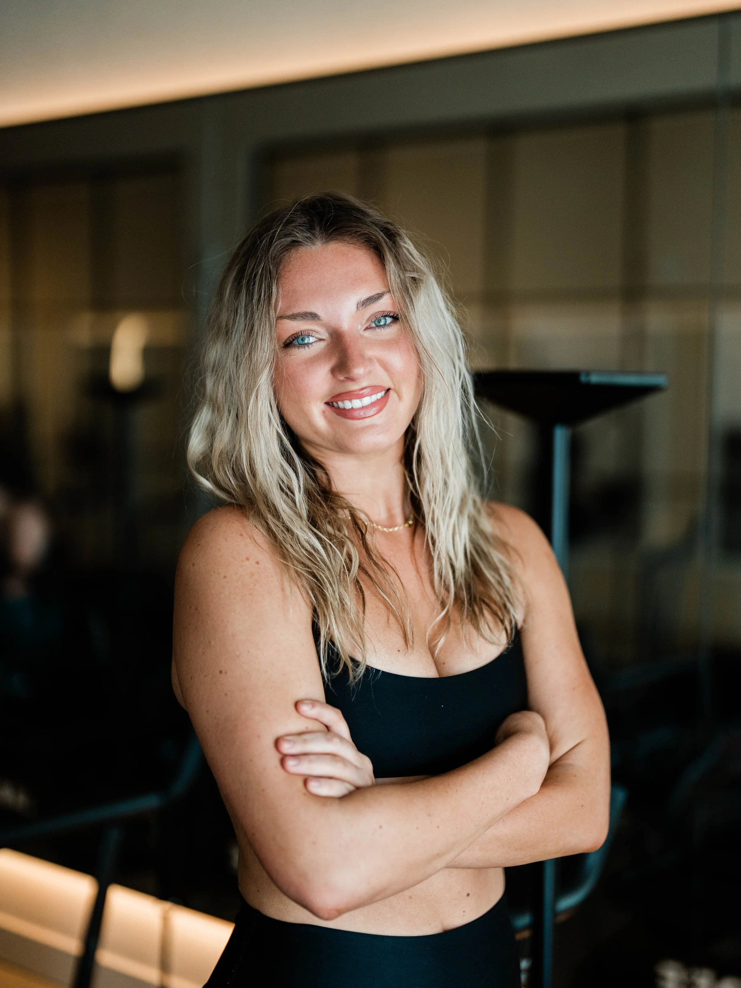 Kylie is a Lagree & VersaClimber instructor with wavy blonde hair and bright blue eyes, smiling with arms crossed, wearing a black spaghetti strap top indoors with a blurred background.