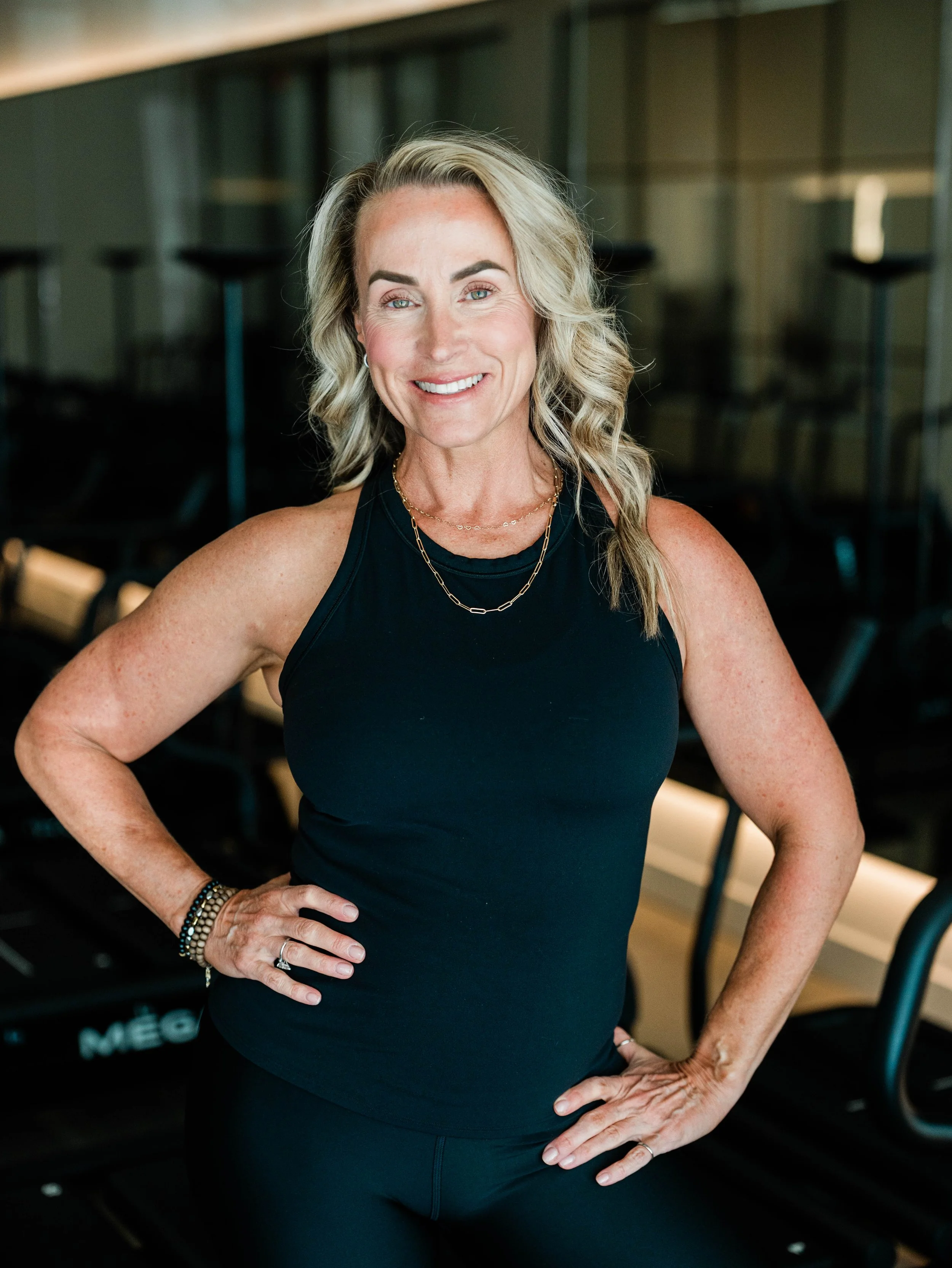 Woman with blonde hair in workout attire smiling and standing with hands on hips in a gym.