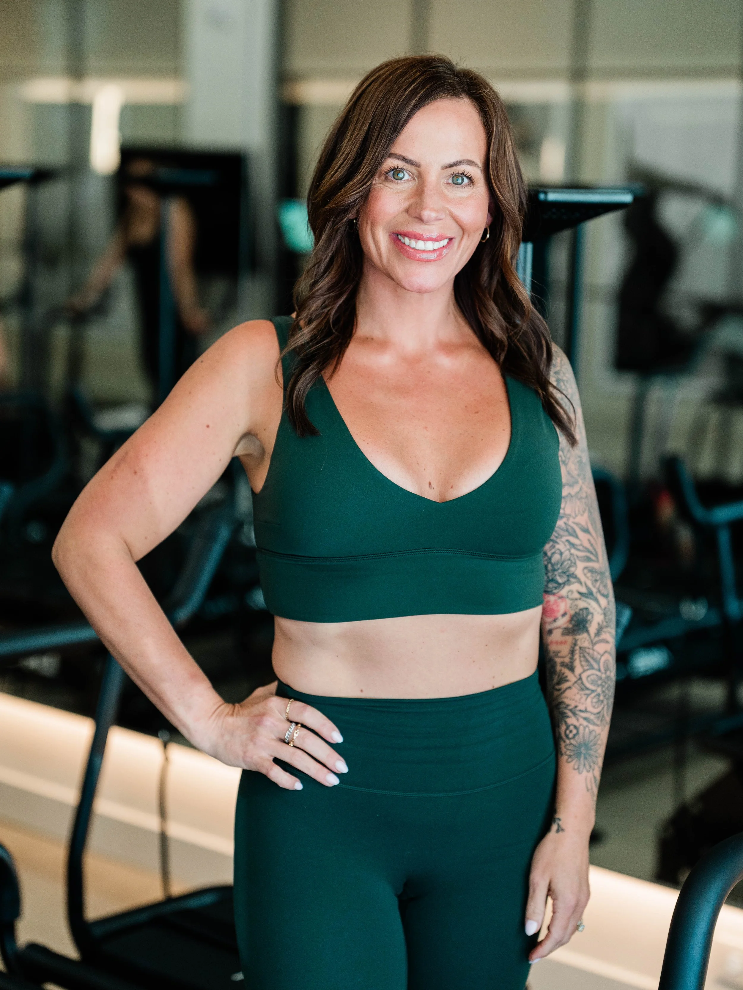 A woman with dark brown hair, blue eyes, and tattoos on her left arm, wearing a dark green sports bra and matching leggings, standing in a gym and smiling at the camera.