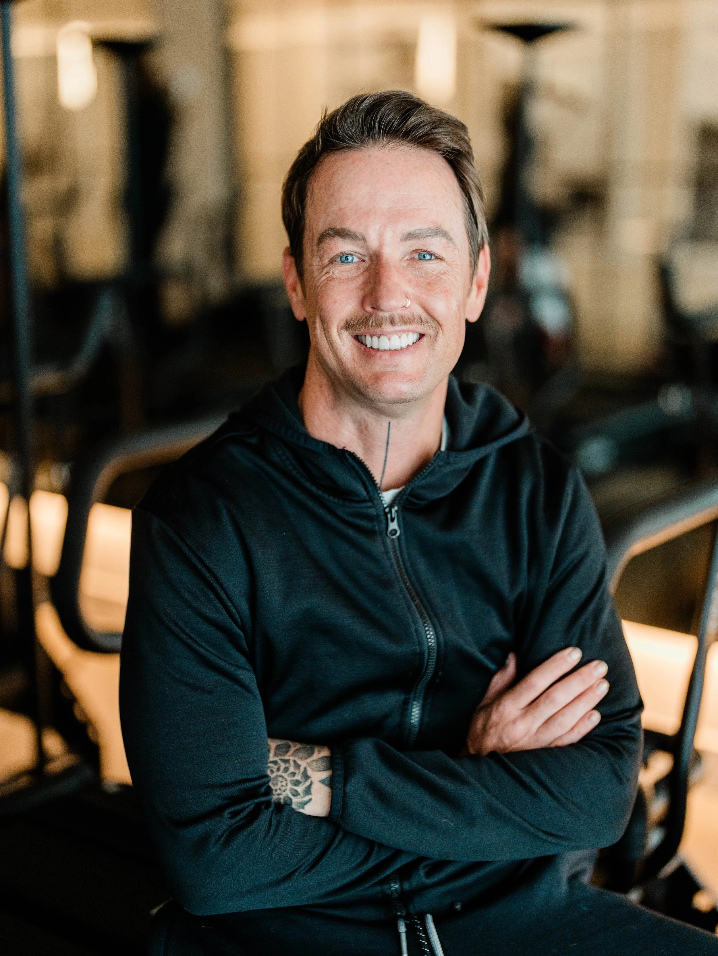 A smiling man with blue eyes, dark hair, and a nose ring, wearing a black zip-up hoodie, sitting with arms crossed in a modern workspace with chairs and tables in the background.