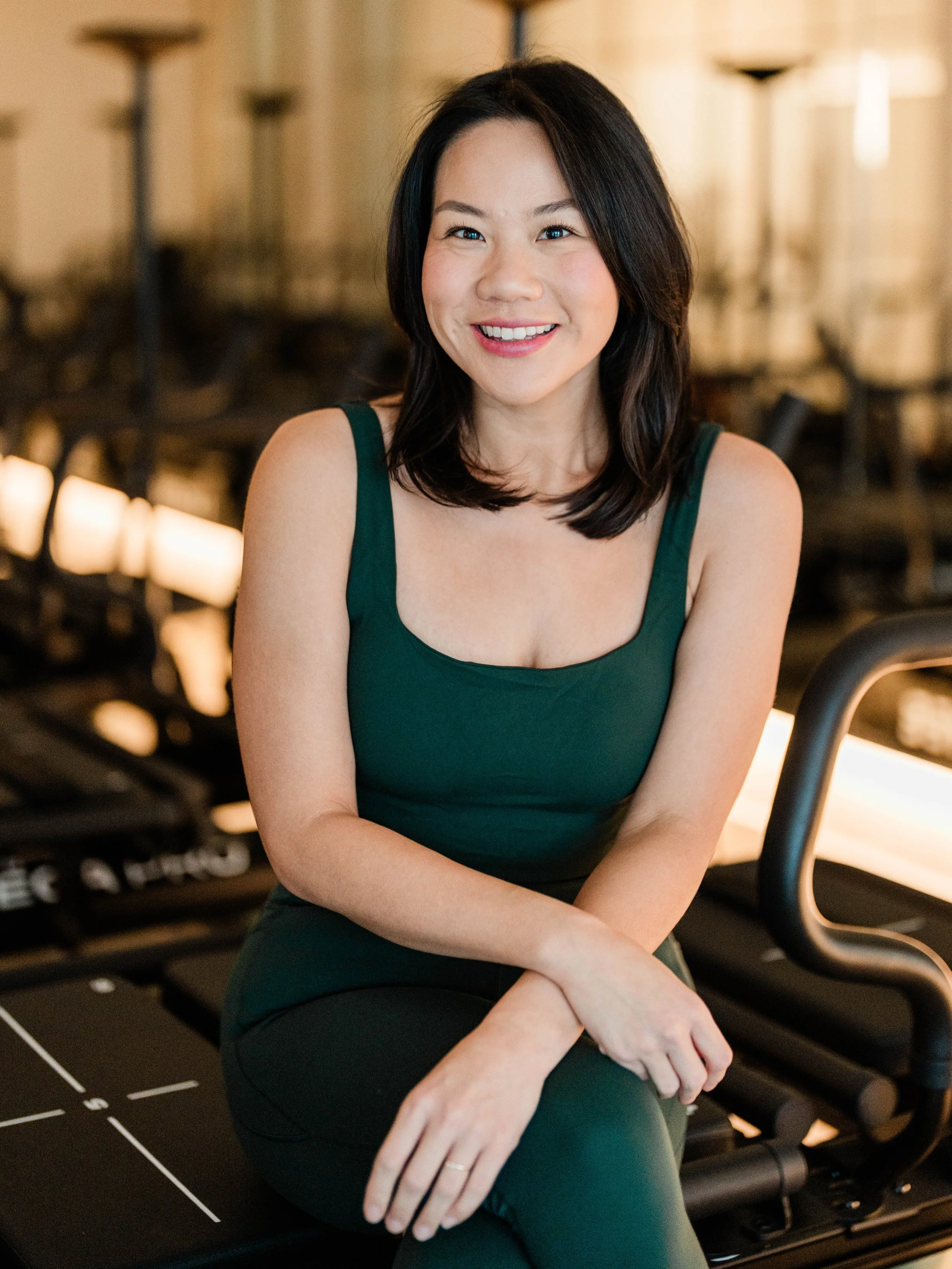 A woman with shoulder-length dark hair, smiling, wearing a dark green sleeveless top, sitting on a black exercise machine in a gym.