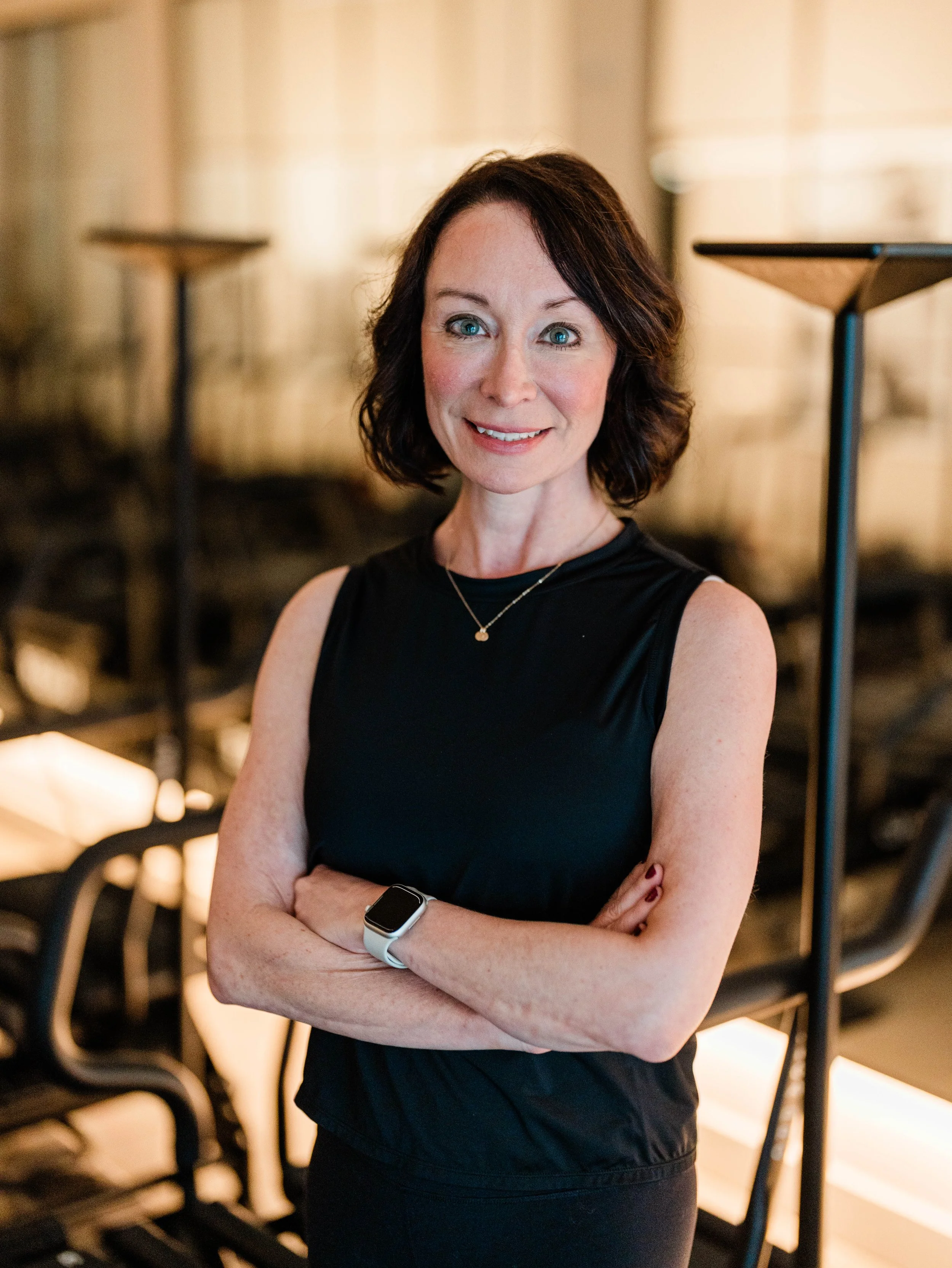 A woman with short dark hair and blue eyes, wearing a sleeveless black top, a necklace, a smartwatch, and maroon nail polish, smiling with arms crossed in a dimly lit indoor setting.