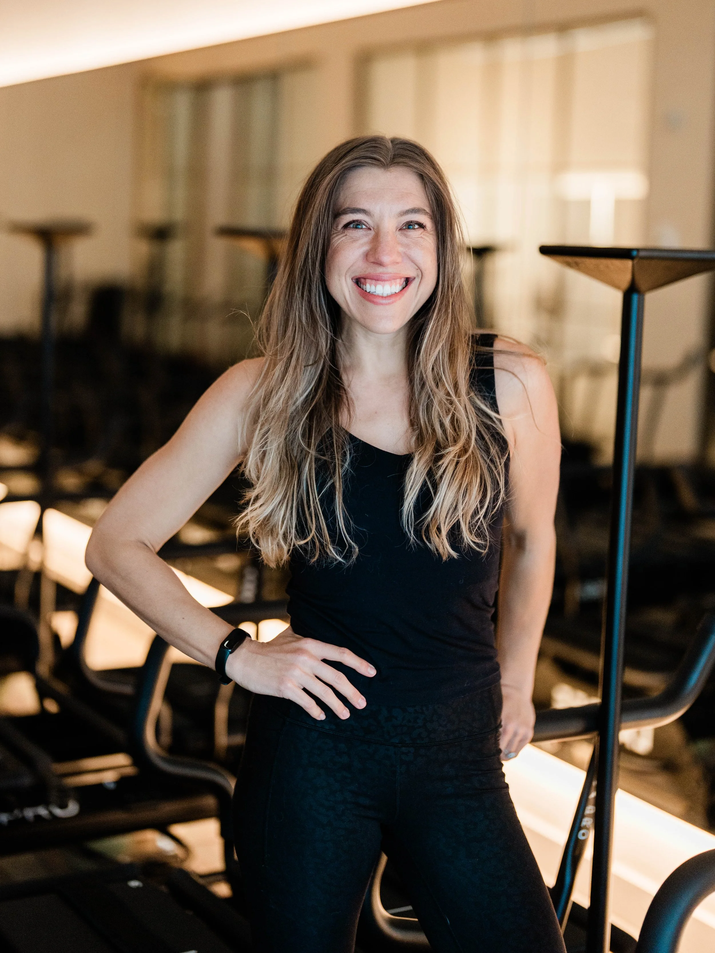A woman with long, wavy blonde hair smiling in a gym or fitness studio, wearing a black sleeveless top and black leggings.