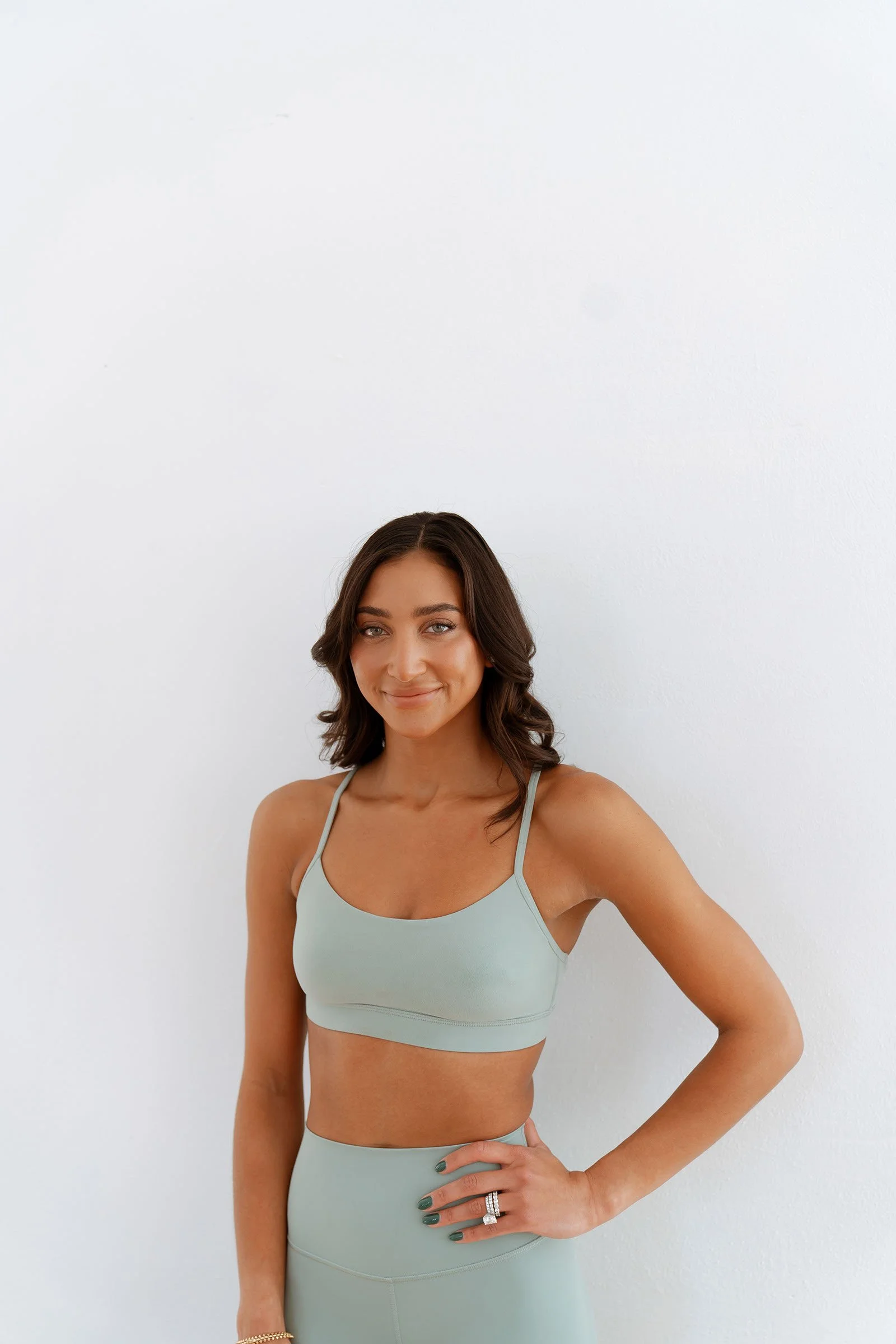 A woman with wavy brown hair, wearing a light gray sports bra and matching leggings, standing against a plain white wall.