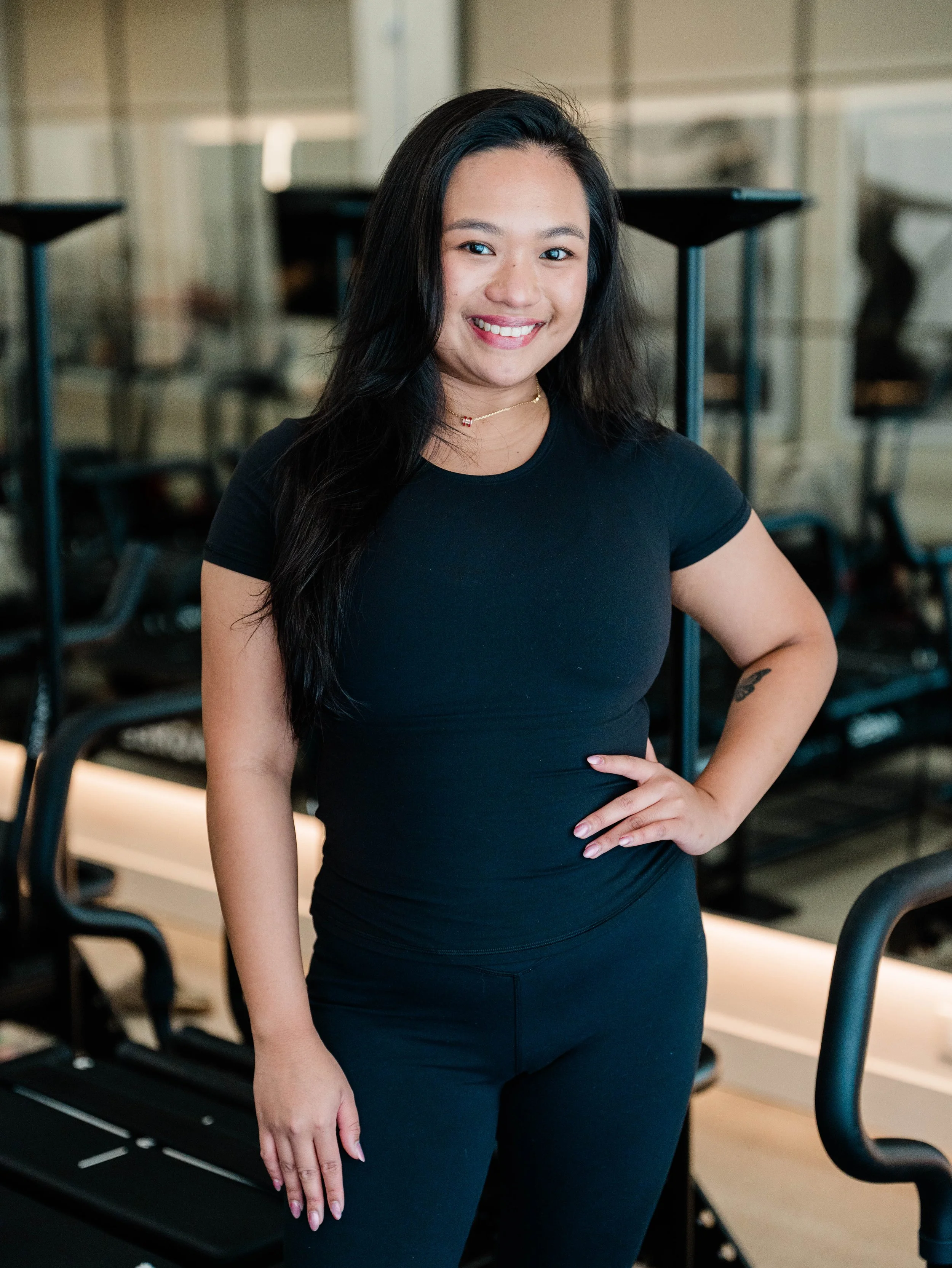 A woman with long black hair wearing a black t-shirt and black pants, smiling and standing in a modern gym or fitness studio.