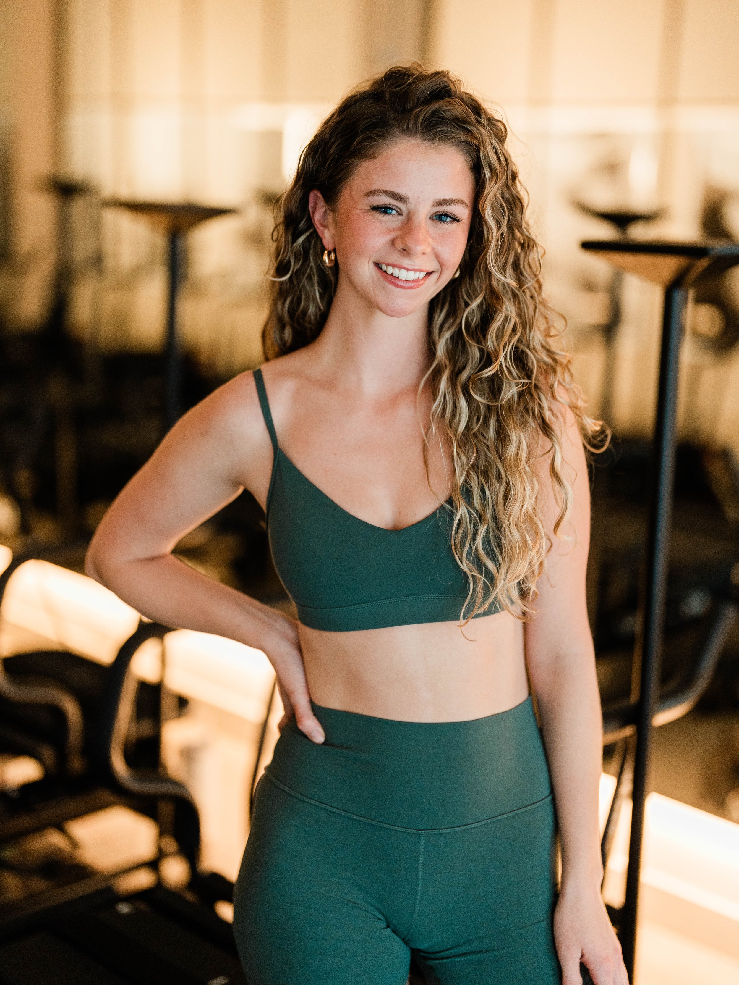A young woman with long, curly blonde hair and fair skin smiling in a gym, wearing a dark green sports bra and matching high-waisted leggings.