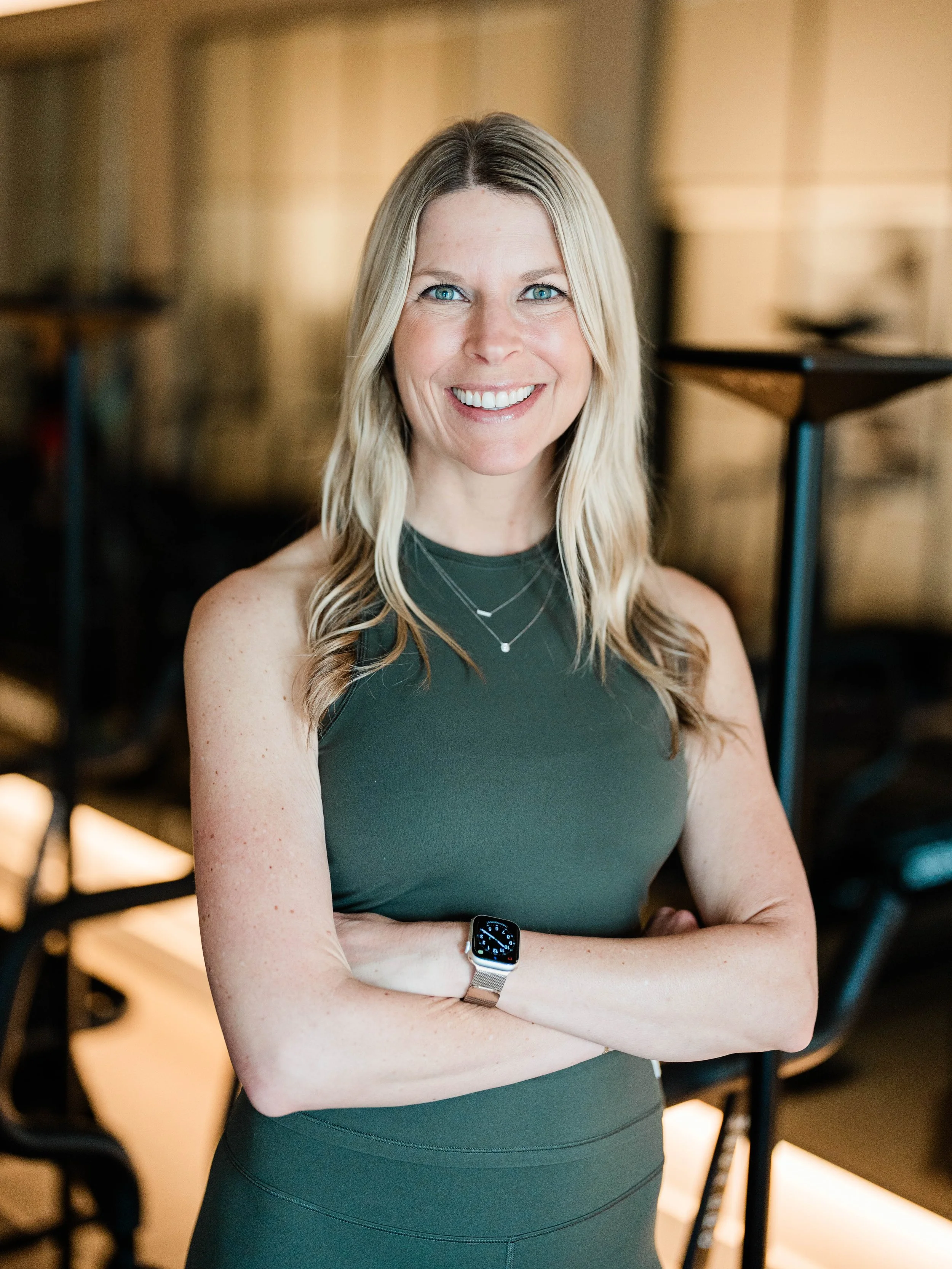 Lindsey is a VersClimber Instructor at Soma Lab. She is pictured wearing a dark green workout top and a smartwatch, standing with arms crossed.