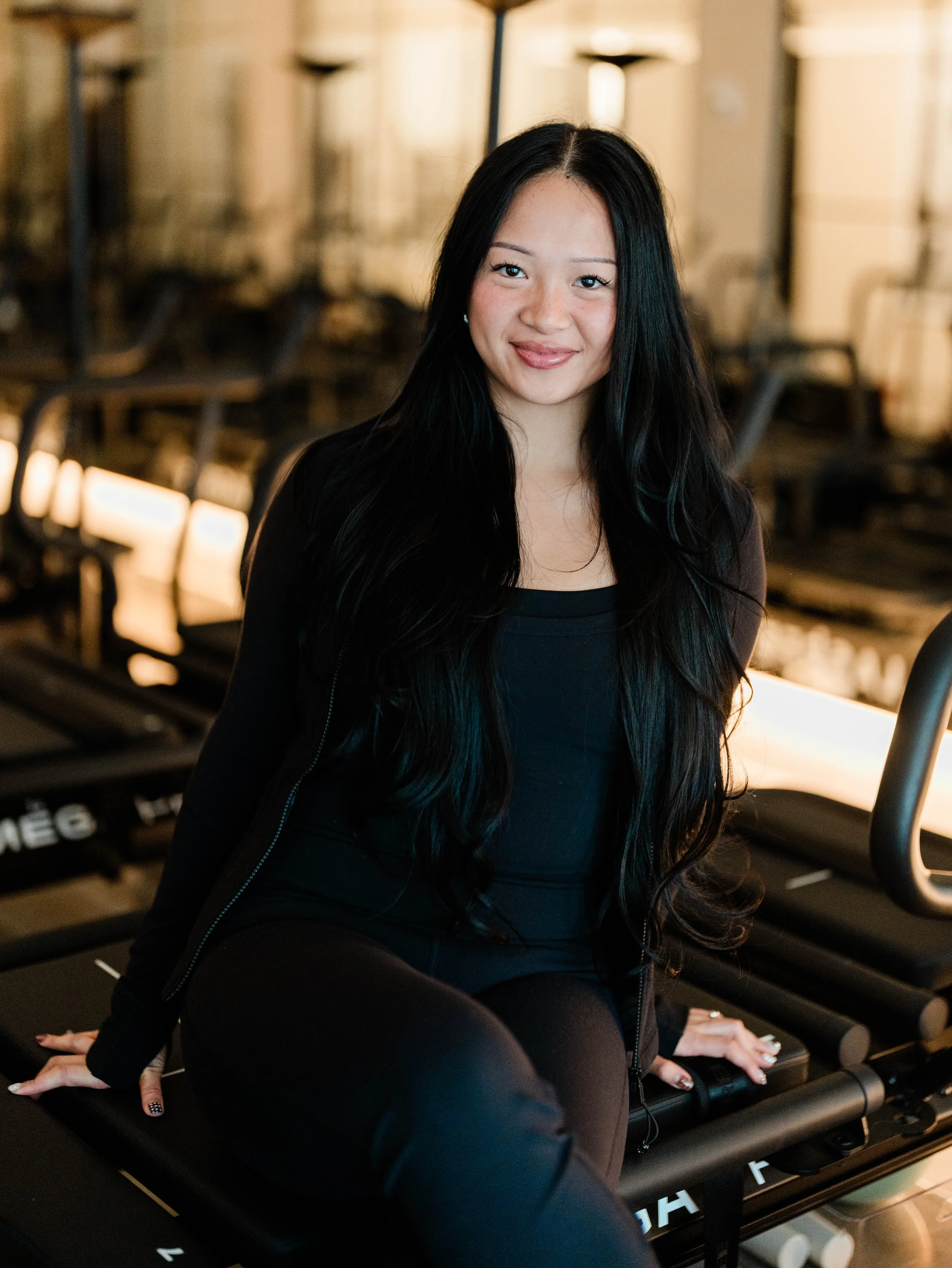 A young woman with long black hair smiling while sitting on a treadmill in a gym.