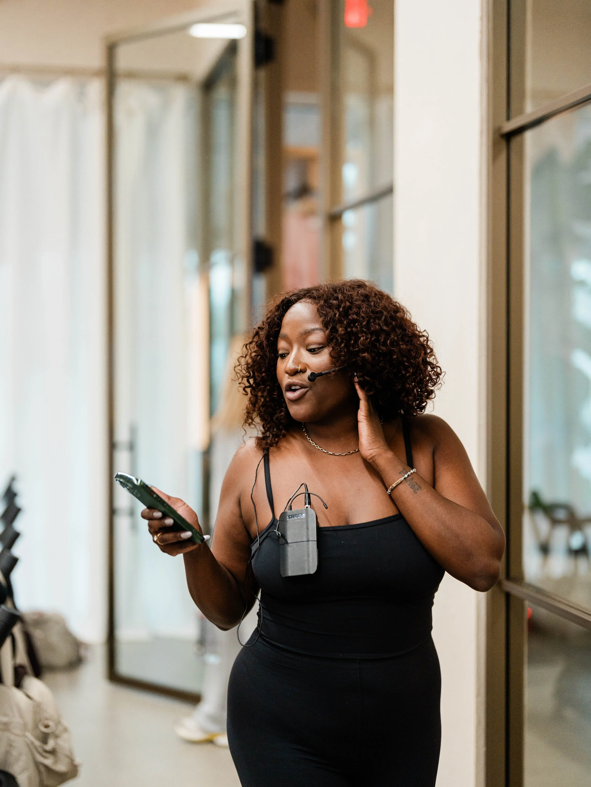 A woman in a black dress with a microphone headset, holding a smartphone and standing indoors near windows.