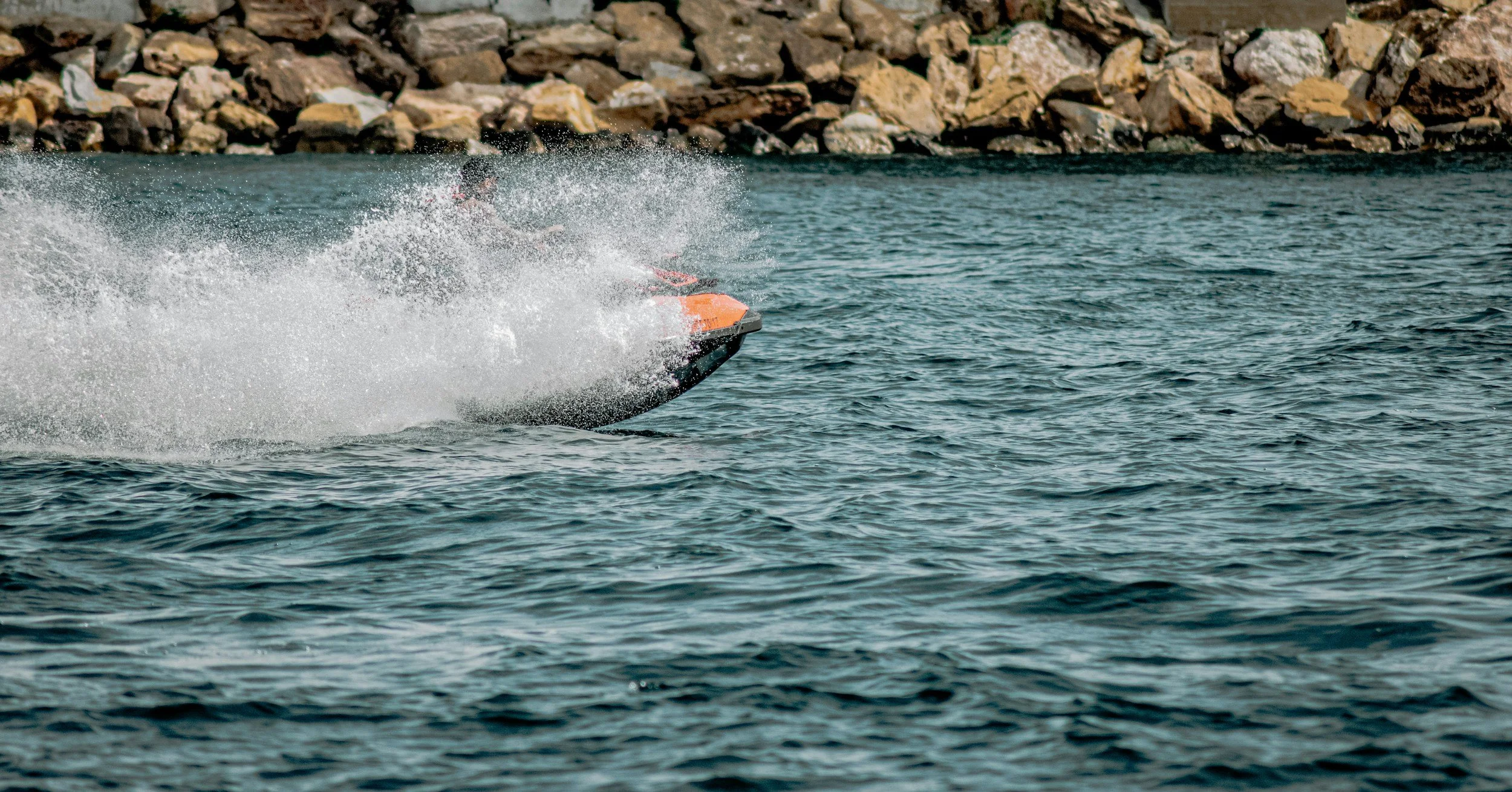 A speedboat pwc jet ski seadoo racing on the water, creating a large splash and wake, with a rocky shoreline in the background.