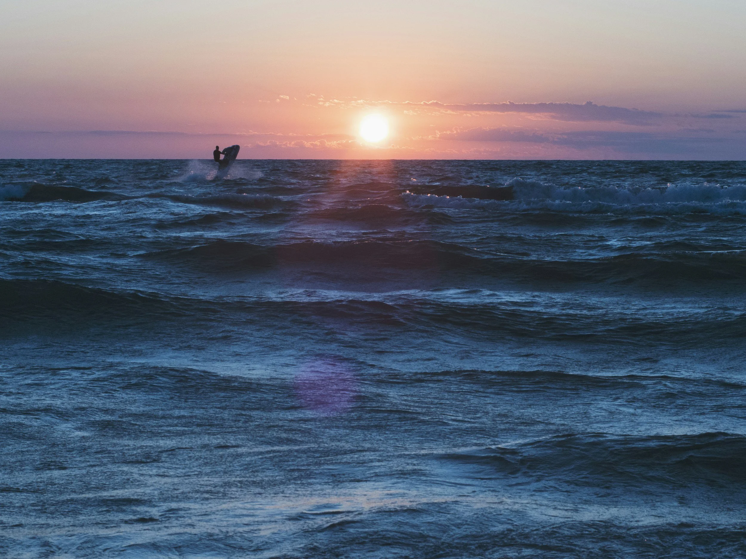 A person riding a jet ski on the ocean during sunset, with the sun low on the horizon and colorful sky in the background.