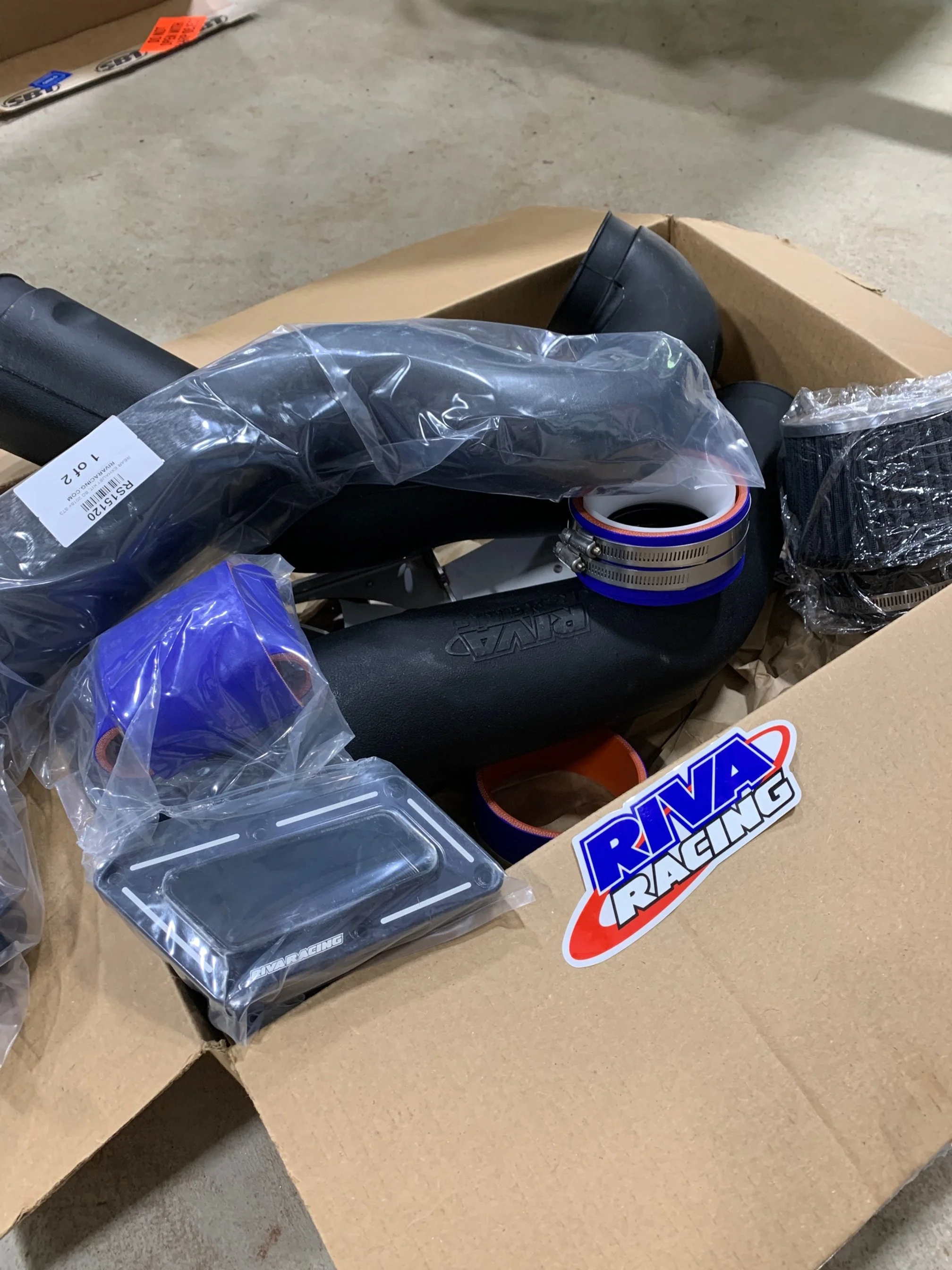 A cardboard box with various racing car parts and accessories, including a steering wheel, clamps, and packaging materials, labeled with a RAV Racing sticker exhaust intake impeller filter