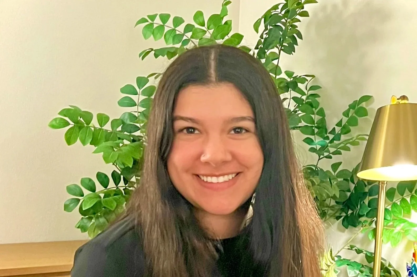 A young woman with long brown hair smiling indoors, standing in front of a large green leafy plant and a gold-colored lamp.
