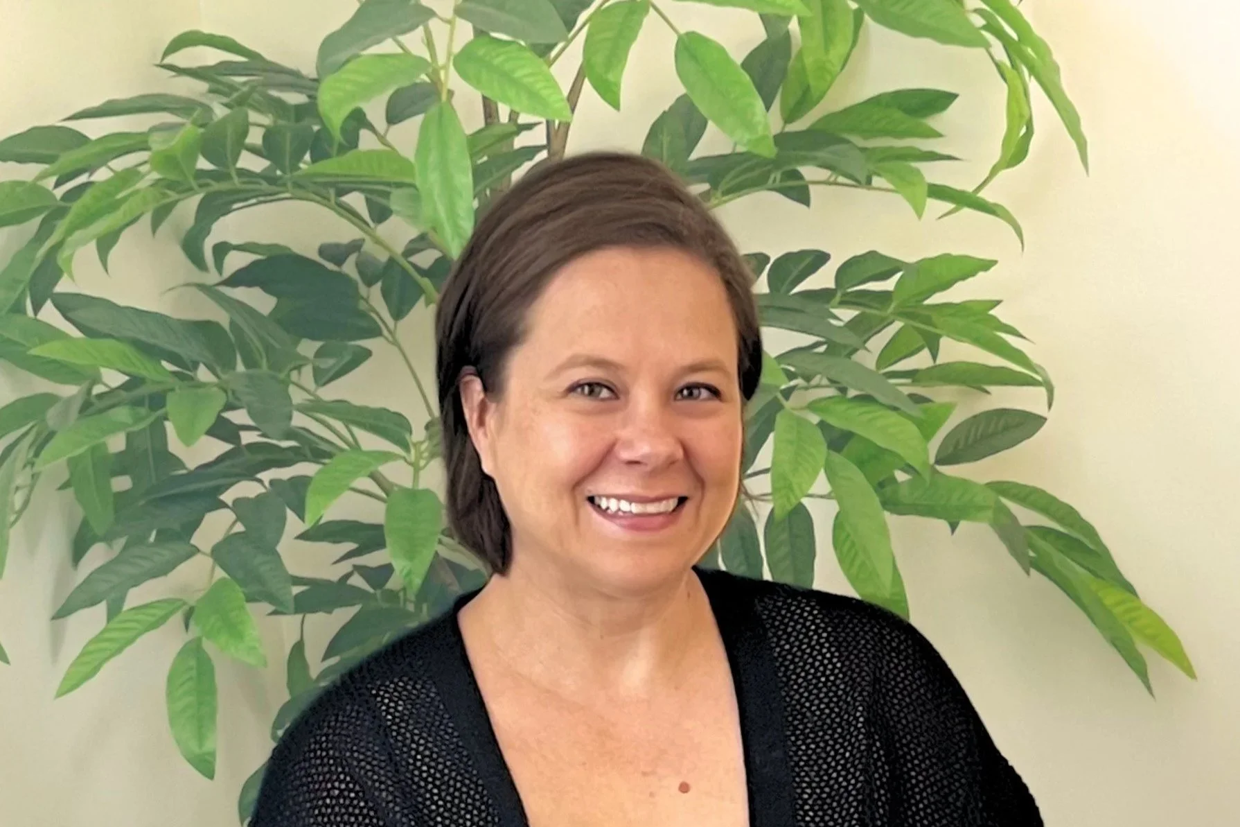 A woman with short brown hair smiling in front of a large green leafy plant.