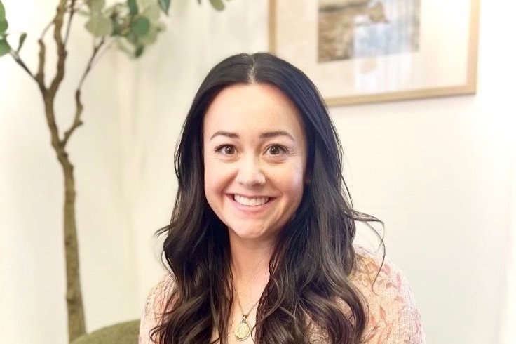 A woman with long dark hair smiling indoors, with a potted plant and framed artwork in the background.