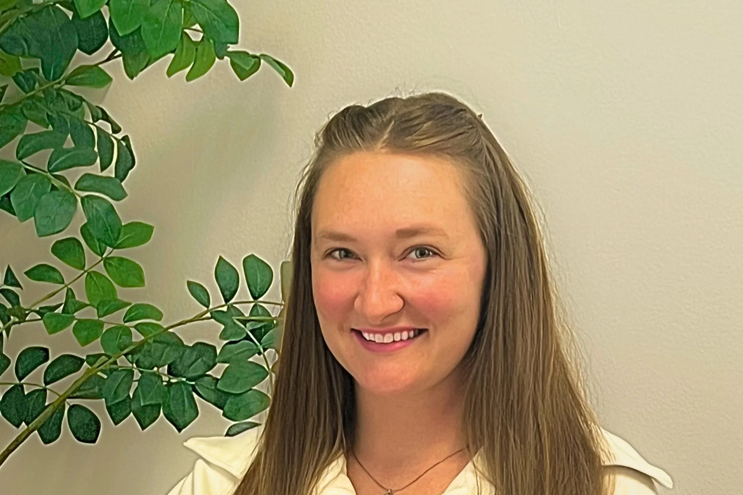 A young woman with long brown hair and a bright smile stands in front of a green leafy plant and a plain beige wall.