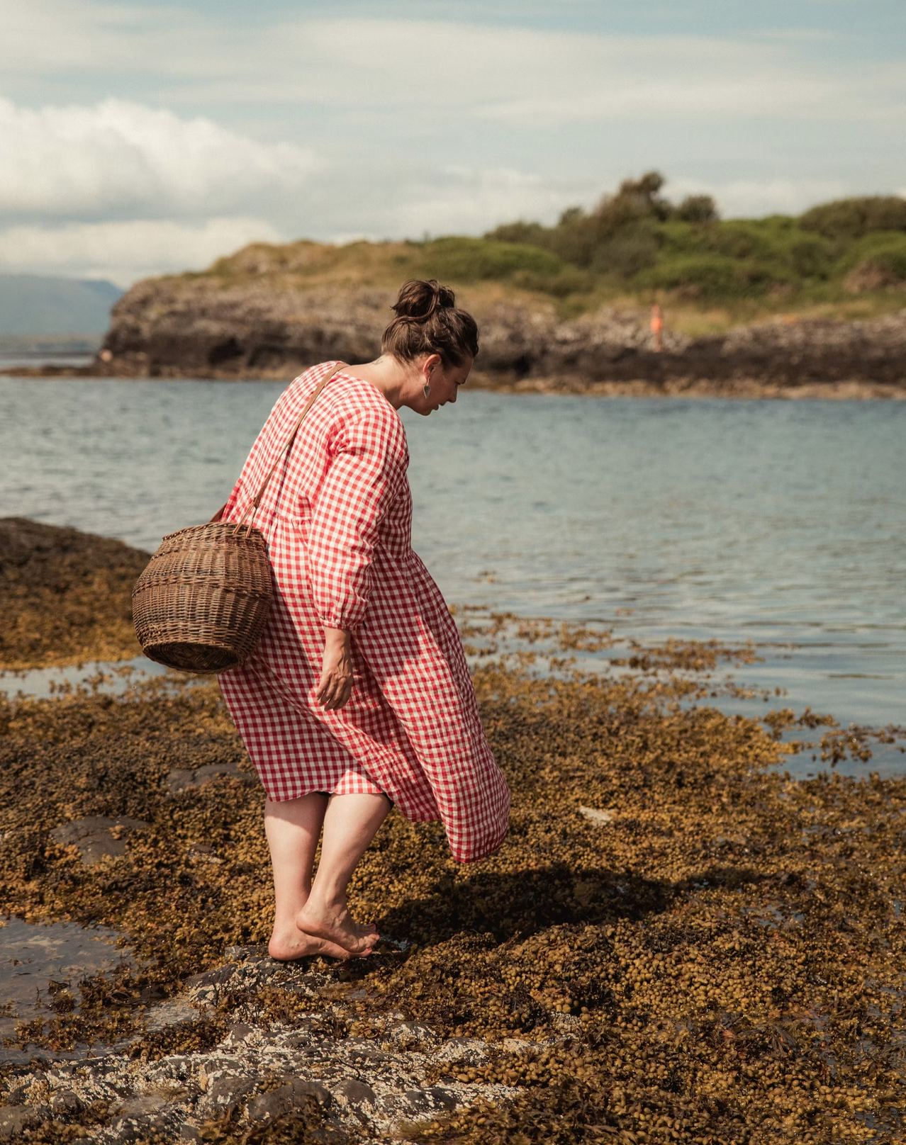 Lucy Cooke in a red and white checkered dress, barefoot, walking on a rocky shoreline covered with seaweed, carrying a wicker basket, with water and a green cove in the background.