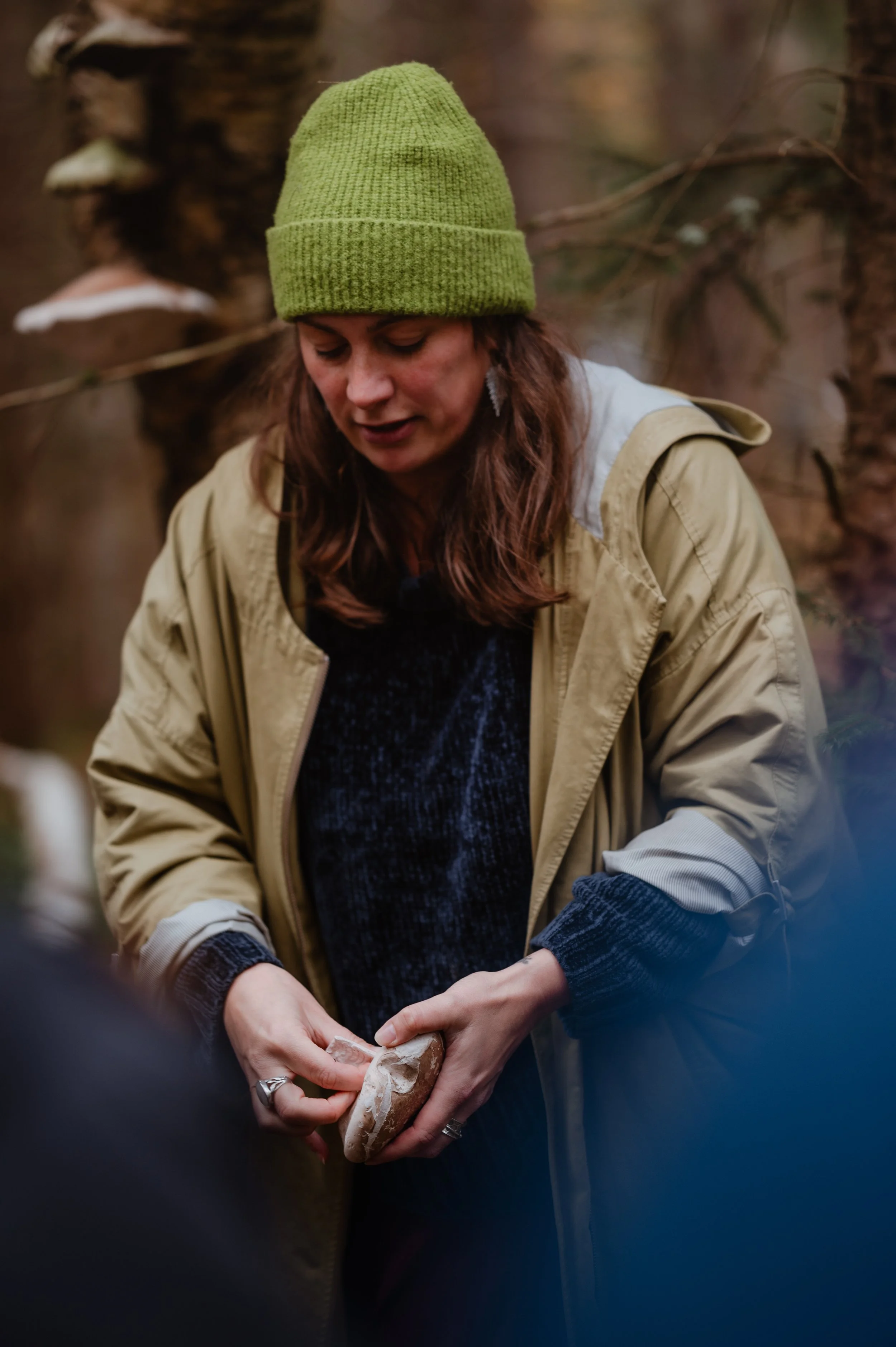 Lucy Cooke, famous forager, a.k.a. the Wild Cooke, wearing a green knit hat and a beige jacket examines a mushroom in a forest.