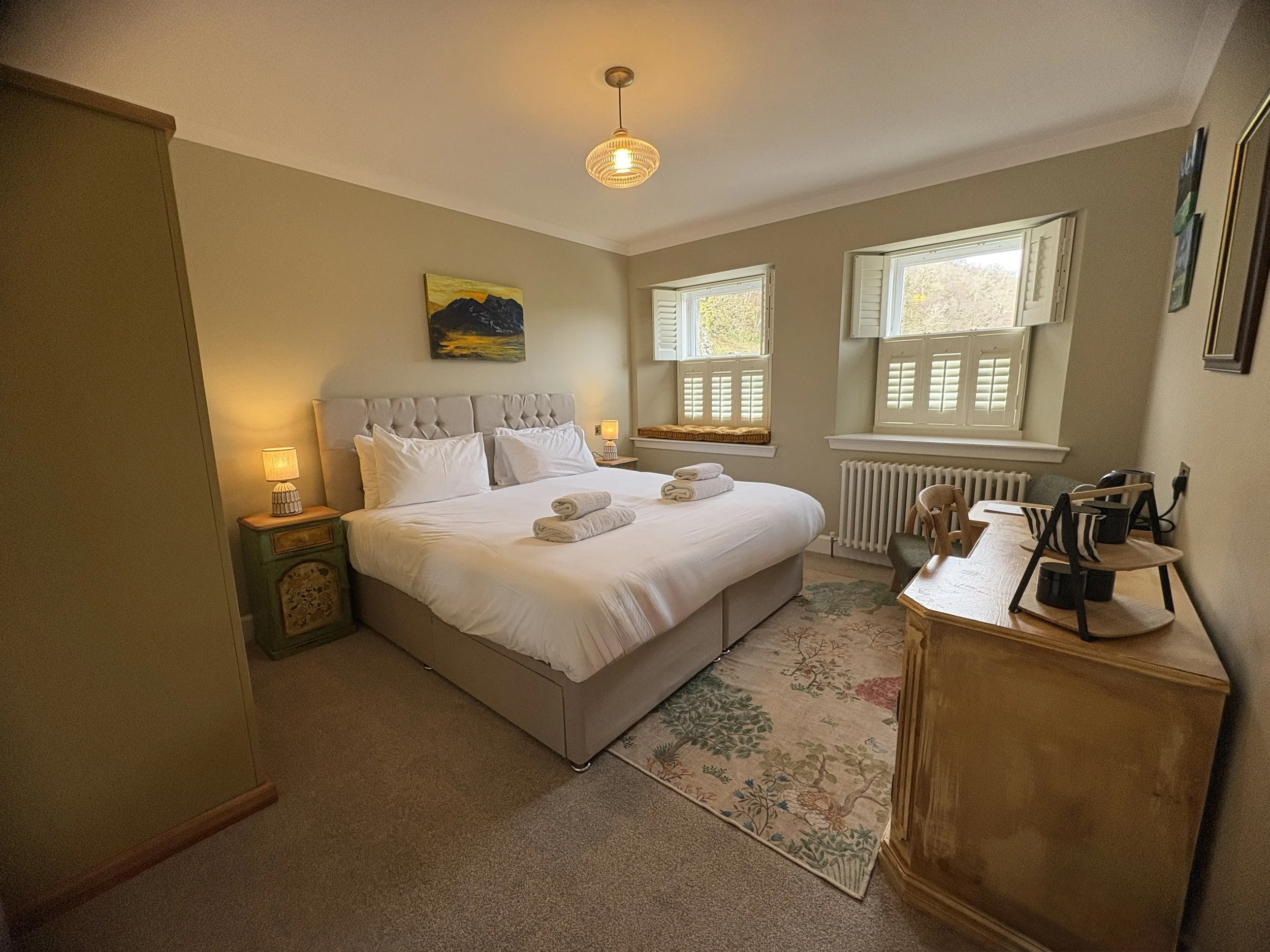 A cozy bedroom with a large bed, two bedside tables with lamps, two rolled towels on the bed, a patterned rug, a window with open shutters, and a vintage-style armchair.