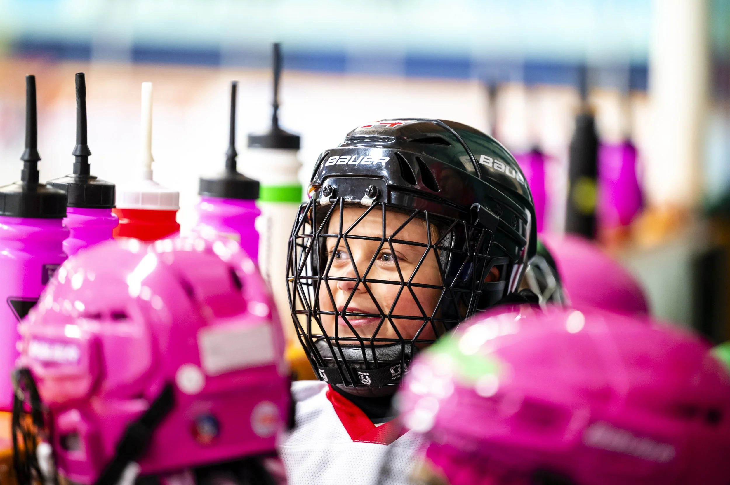 A young hockey player wearing a black helmet with a cage, smiling among pink water bottles in a locker room.