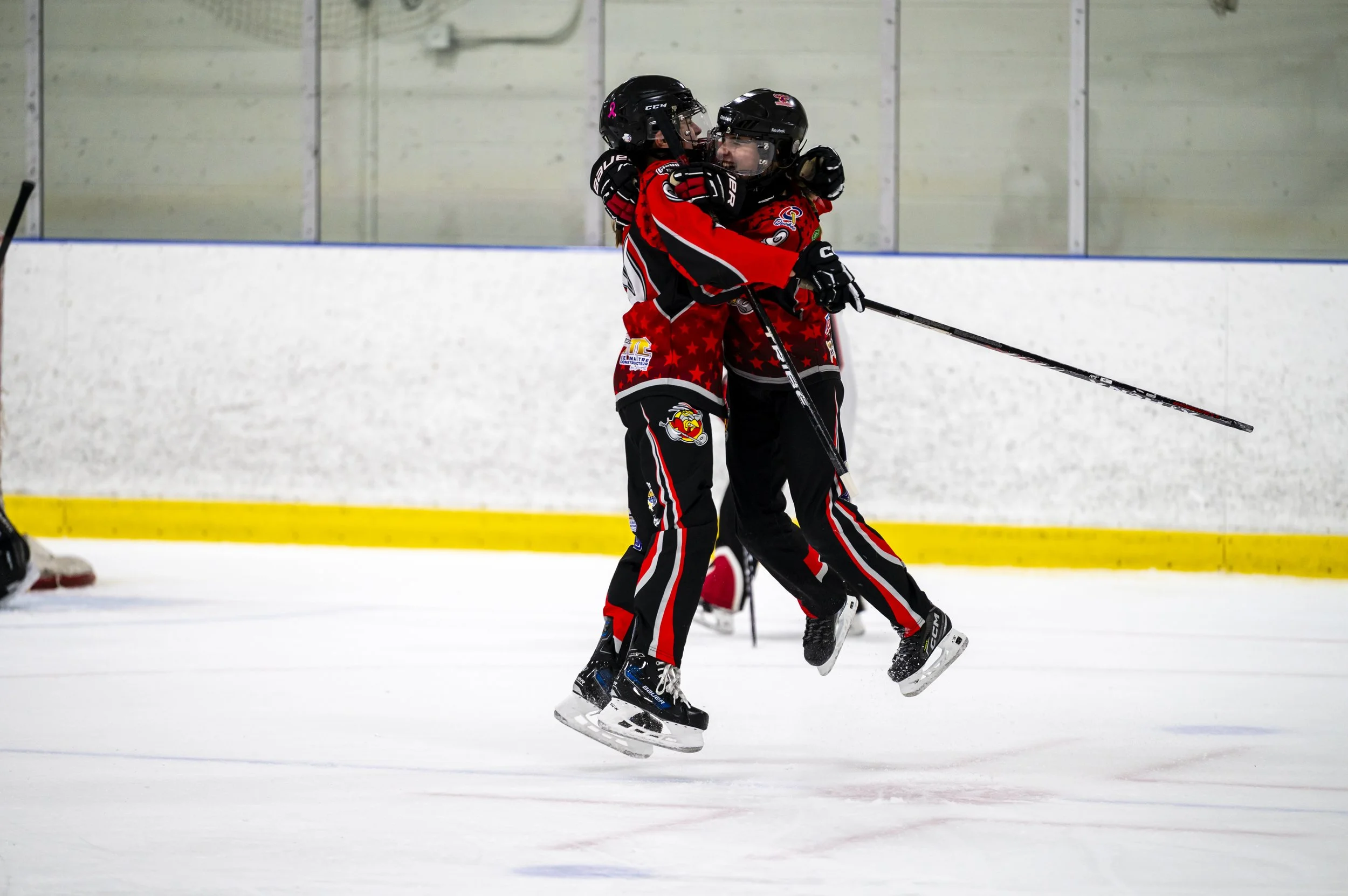 Two ice hockey players in red and black uniforms are celebrating by hugging on the ice rink.