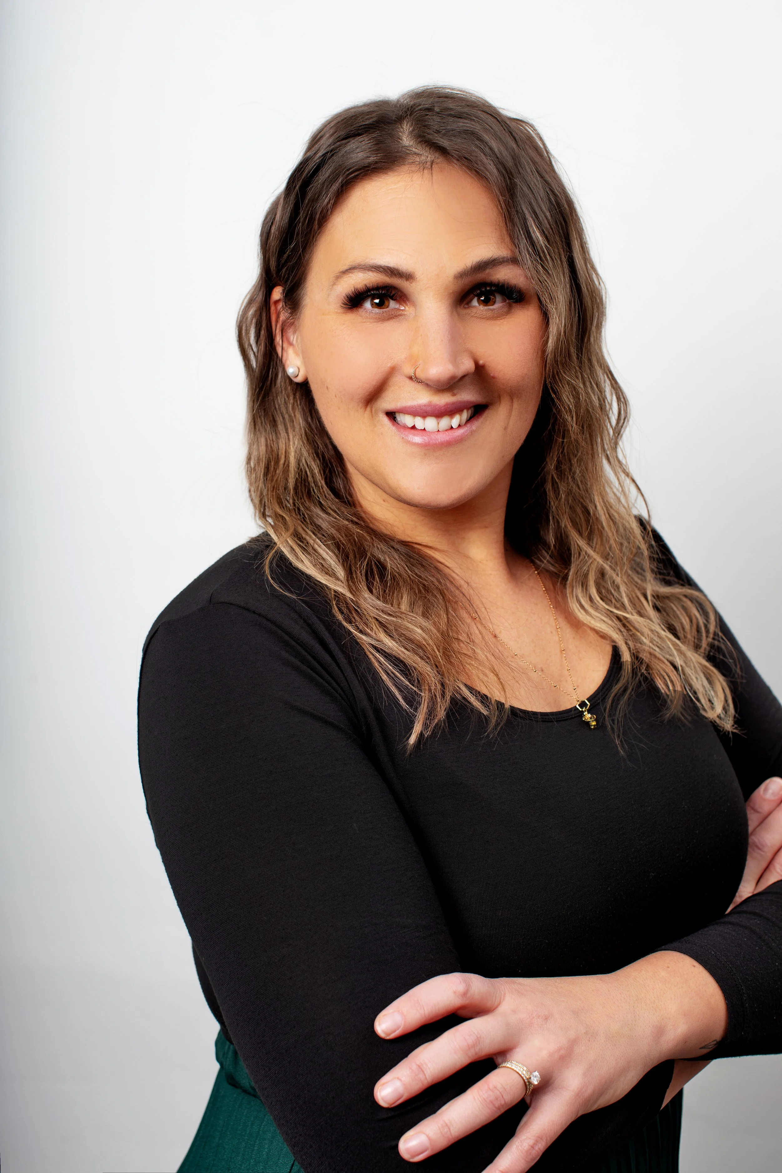 A woman with long wavy hair, wearing a black top, earrings, and a necklace, smiling and crossing her arms in front of a plain white background.