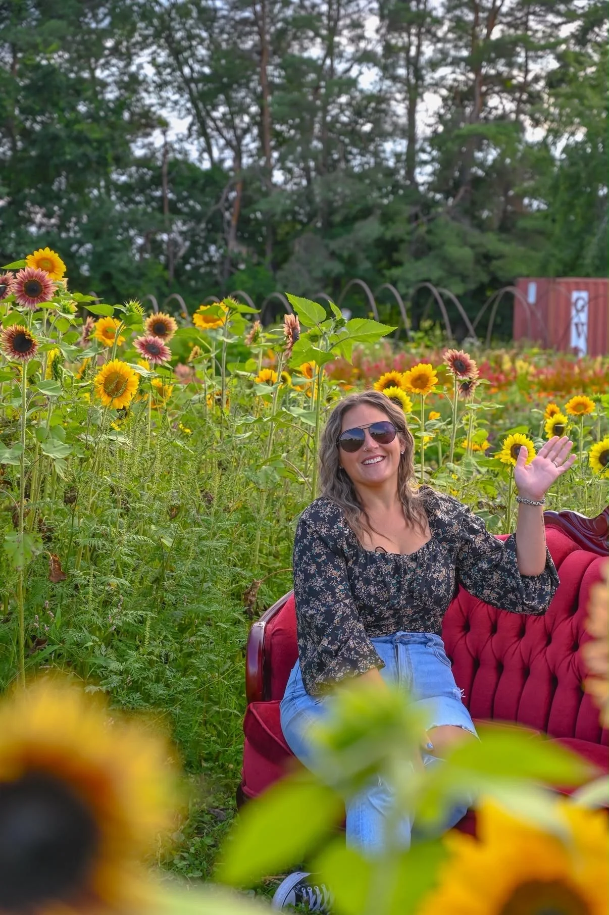 A woman with sunglasses and a floral blouse sitting on a red vintage sofa in a sunflower field, smiling and waving.