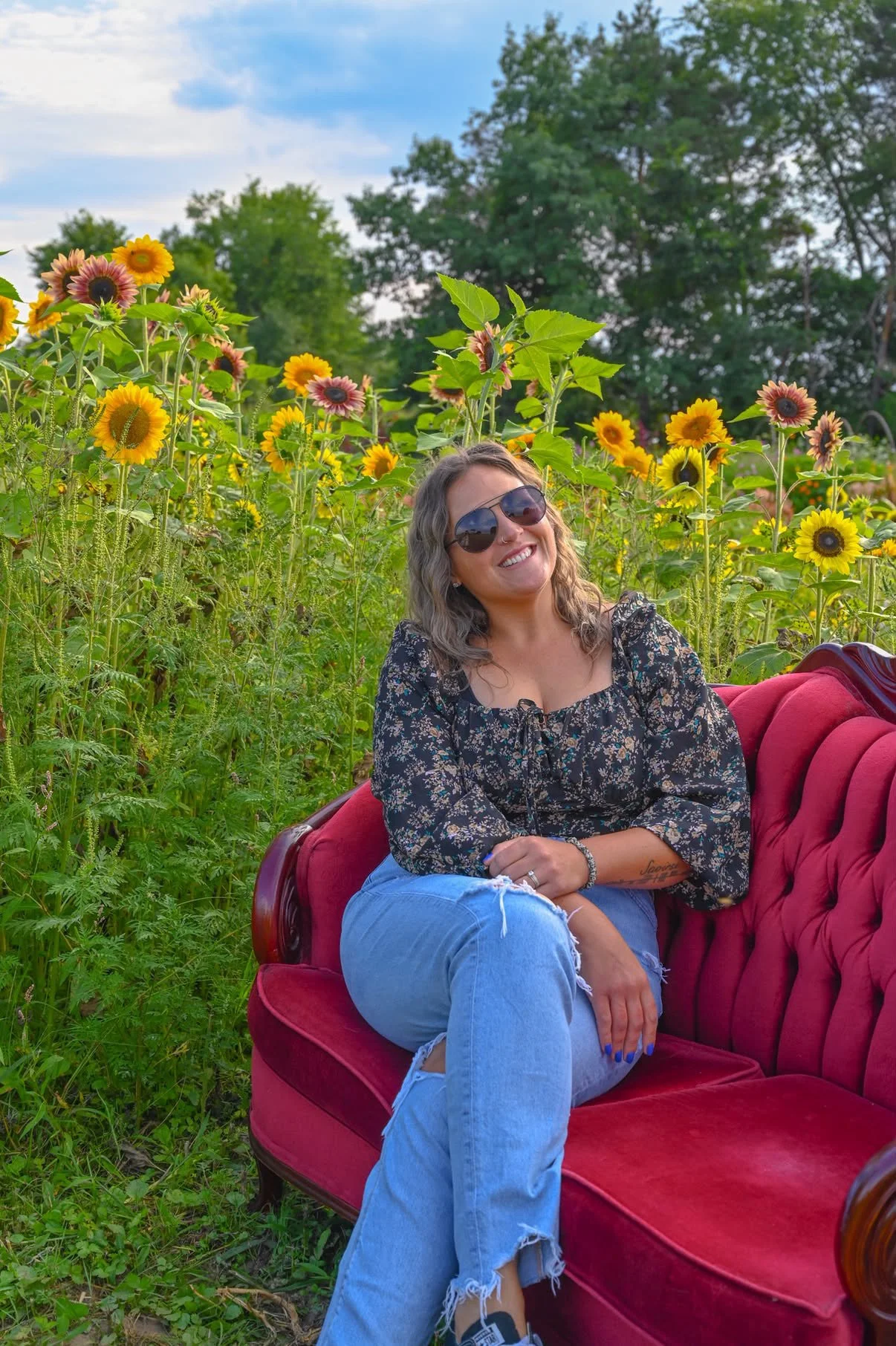 Woman sitting on a red vintage sofa outdoors surrounded by tall sunflowers and green trees, smiling, wearing sunglasses, a dark floral blouse, and ripped blue jeans.