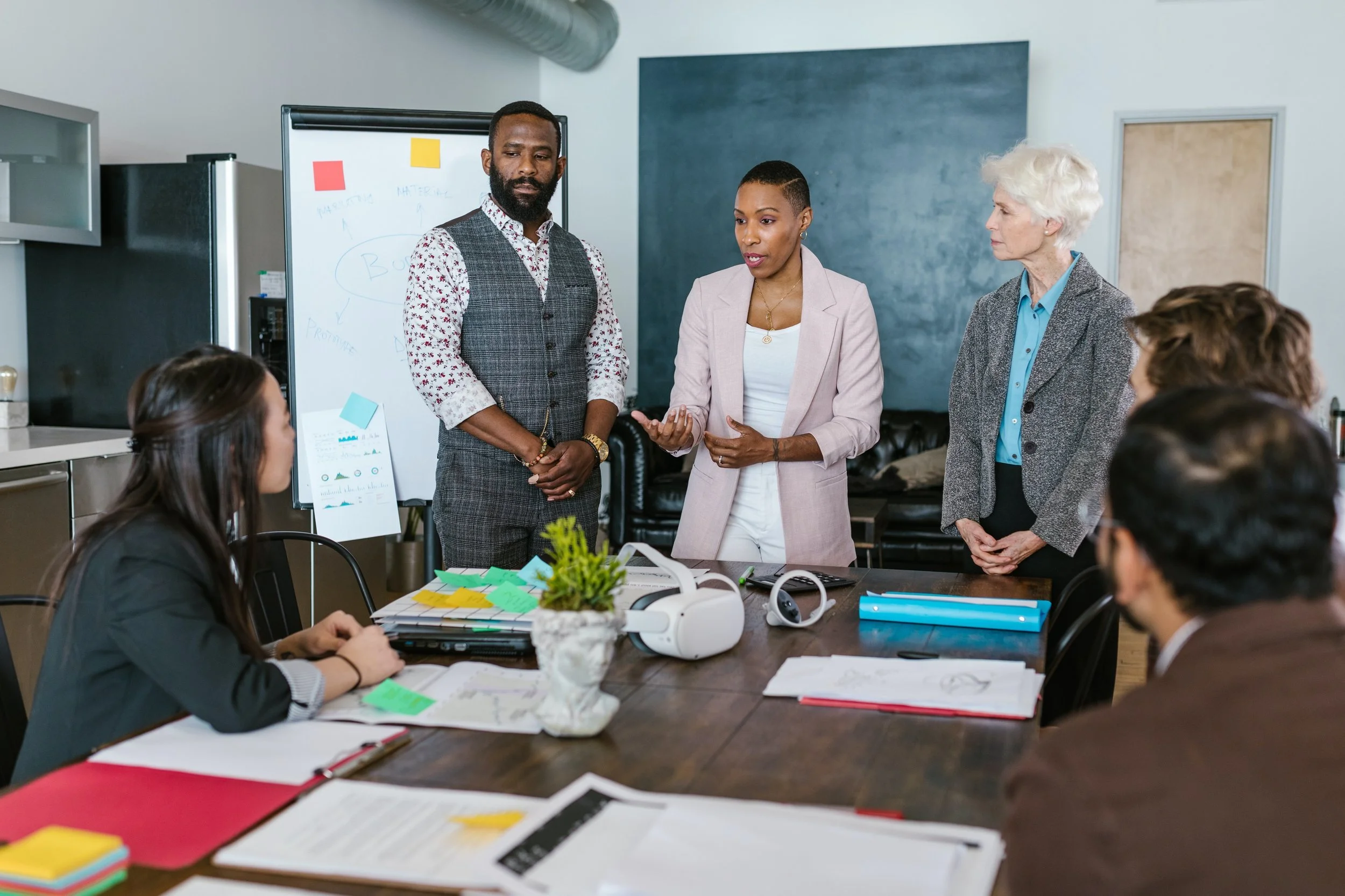 Business meeting with diverse group of people in a conference room, standing and sitting around a table with documents and electronic devices.