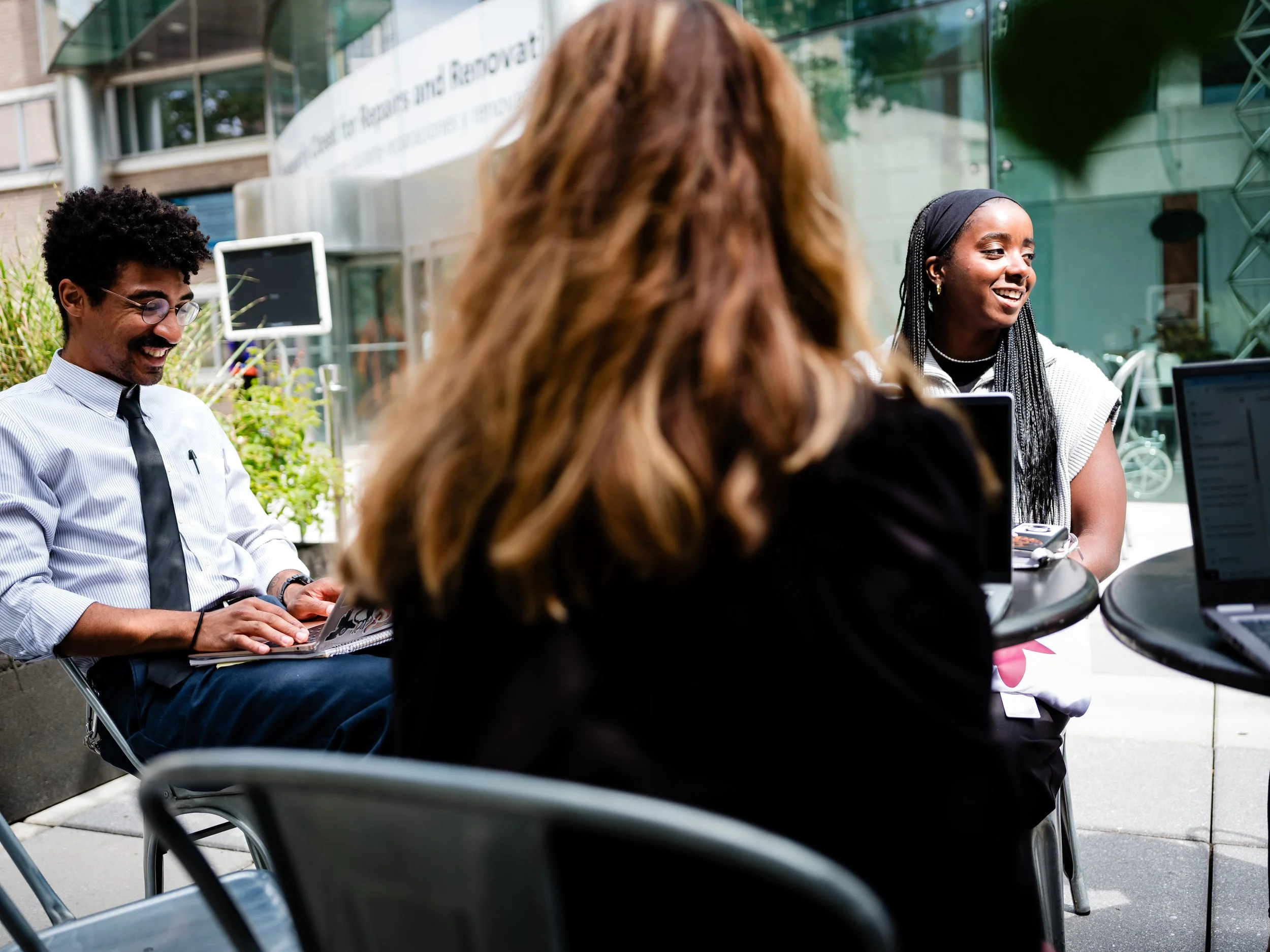 Three people sitting outside at round tables with laptops, engaged in a conversation, smiling, in an urban setting