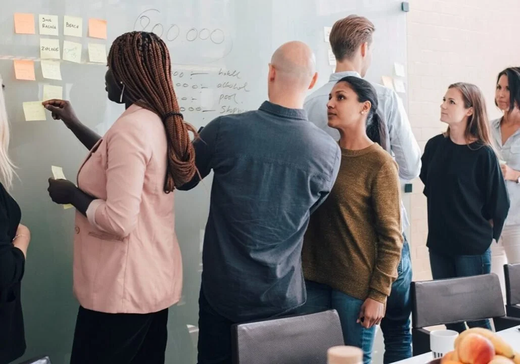 Group of people standing and talking in a meeting room with sticky notes on a whiteboard.