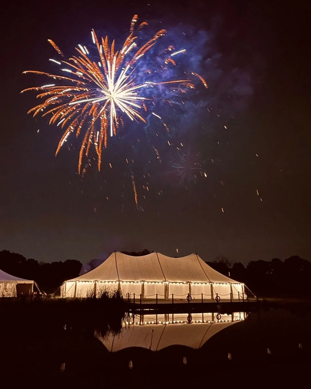Colorful fireworks burst above a warmly lit event tent reflected in a pond at night.