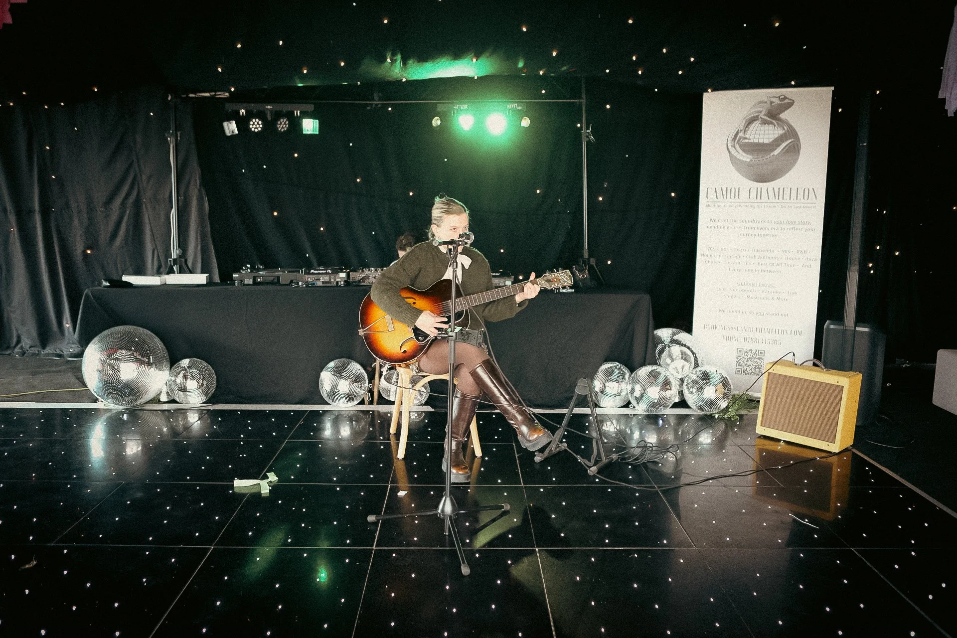 Musician performing with an acoustic guitar and microphone on a star-lit stage with disco balls and DJ equipment.