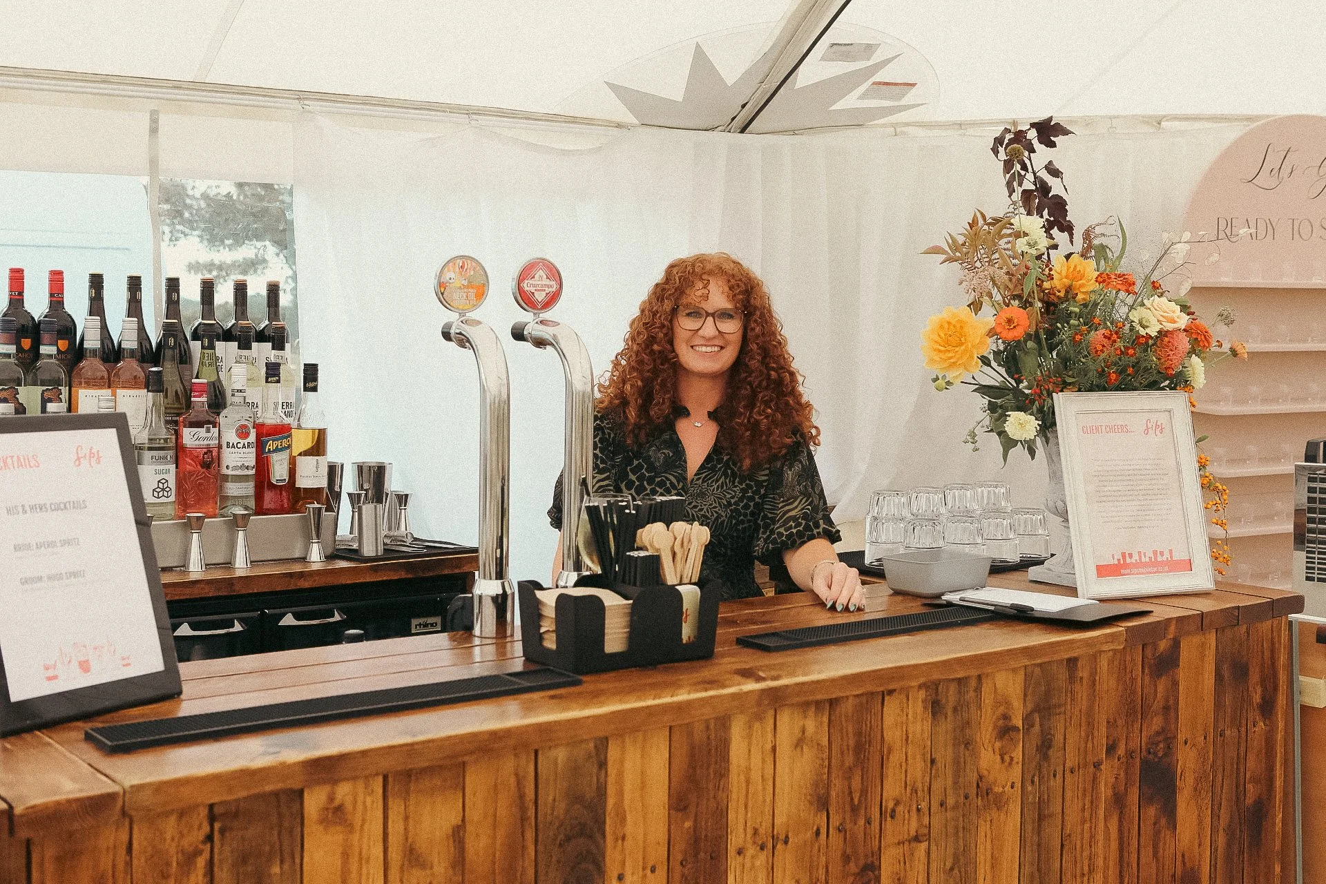 Smiling bartender behind a rustic wooden bar with liquor bottles, beer taps, glassware, and colorful flowers