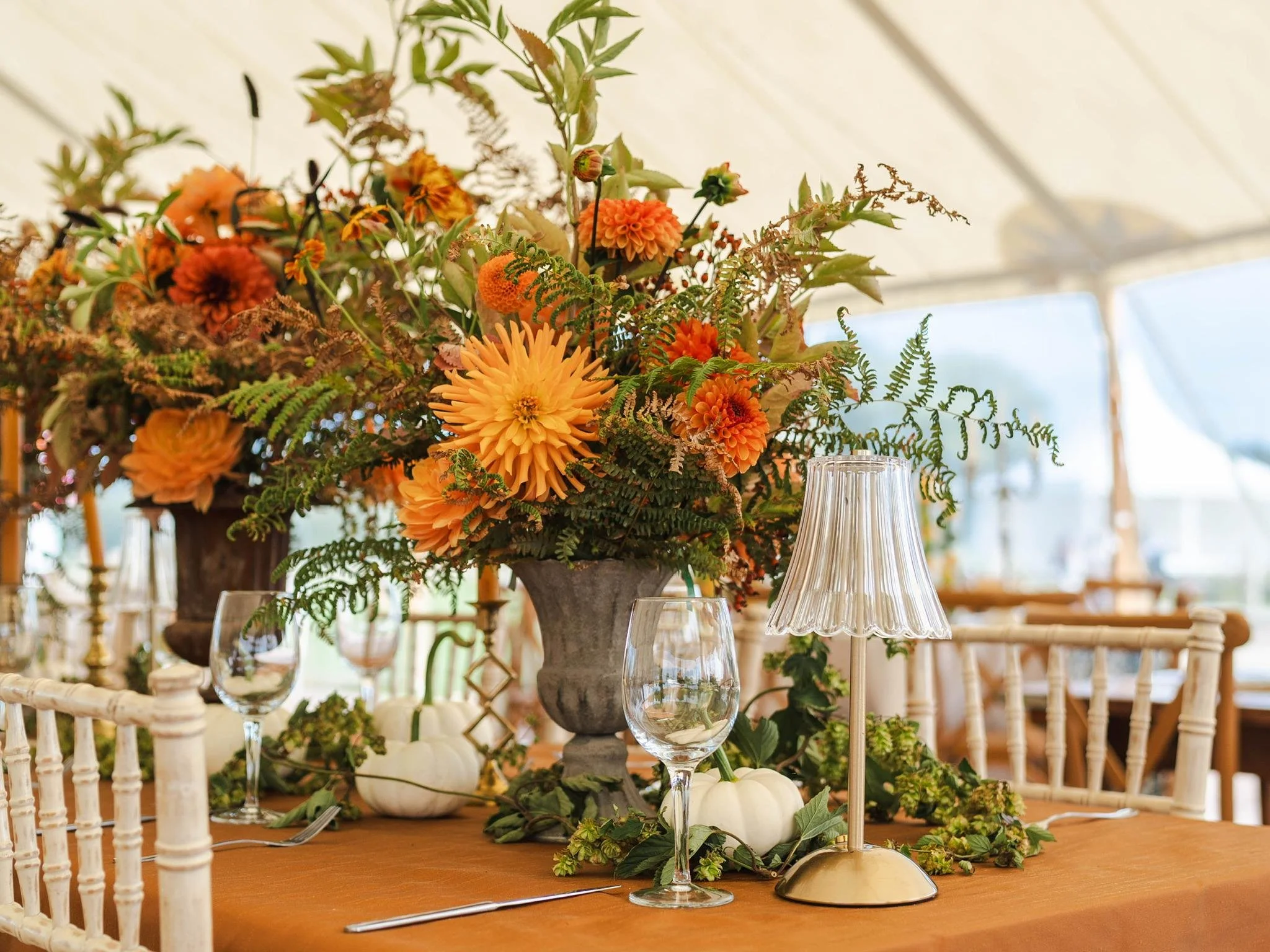 Elegant autumn wedding table centerpiece with orange dahlias, greenery, white pumpkins, glassware, and a table lamp