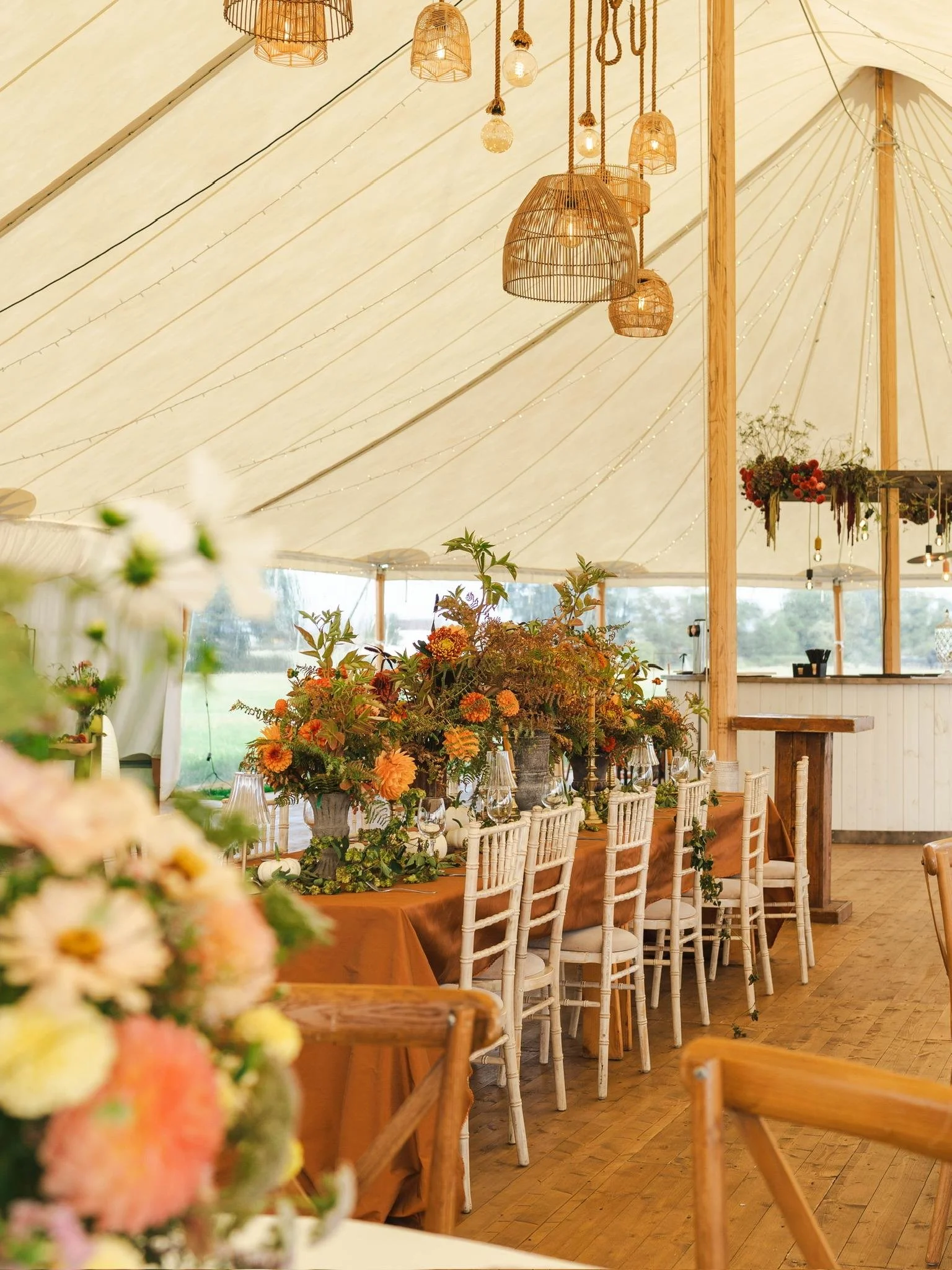 The interior of a traditional pole marquee decorated with autumnal florals, limewash chiavari chairs and rattan feature lighting