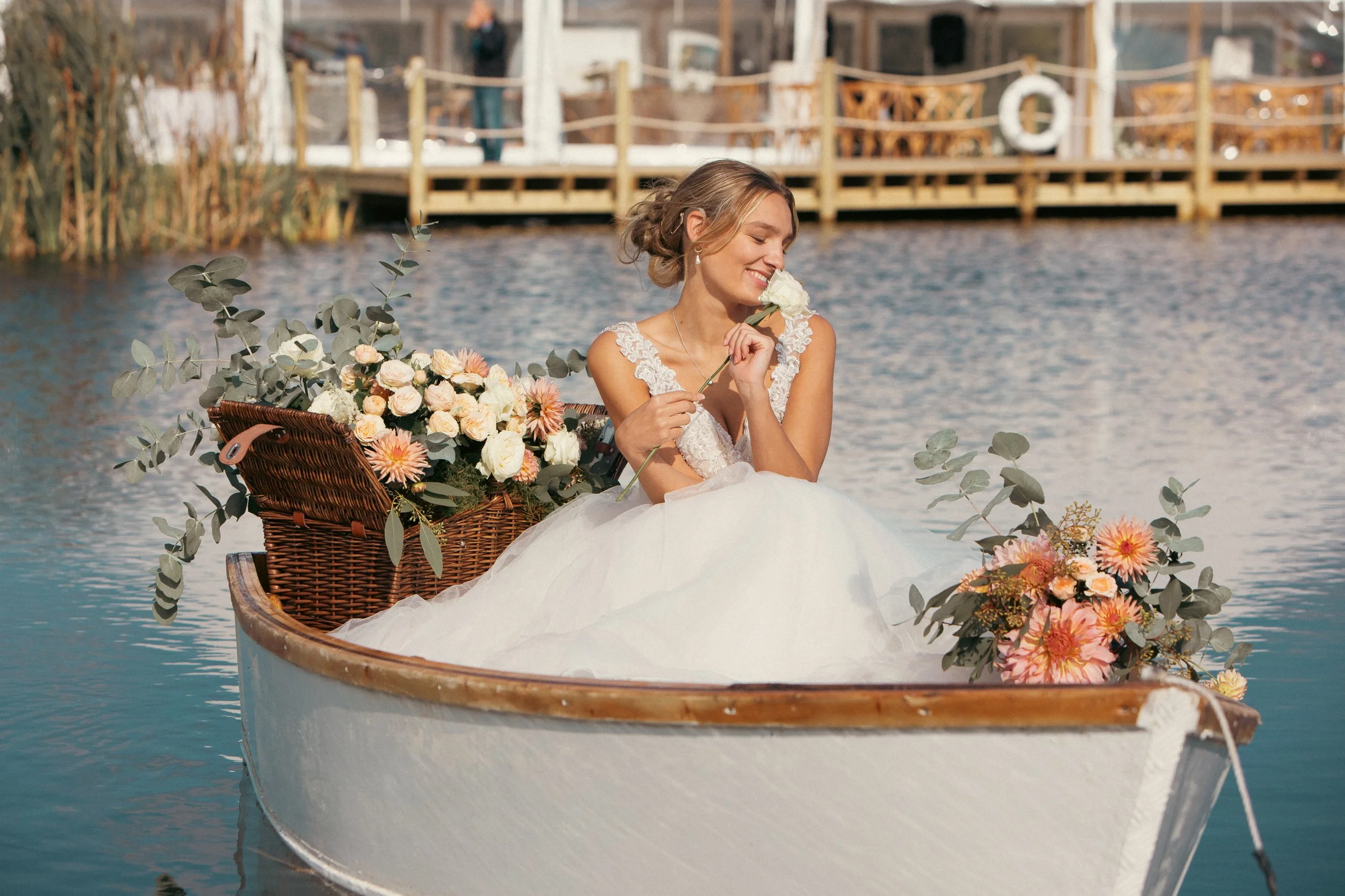 Smiling bride in a white lace wedding dress holding a rose on a flower-decorated boat.