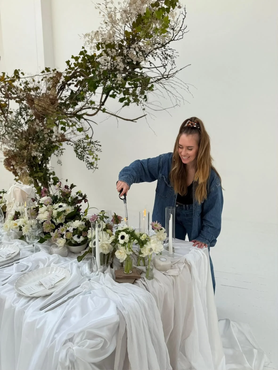 Woman arranging flowers and candles on an elegant white-draped dinner table beneath a floral arch