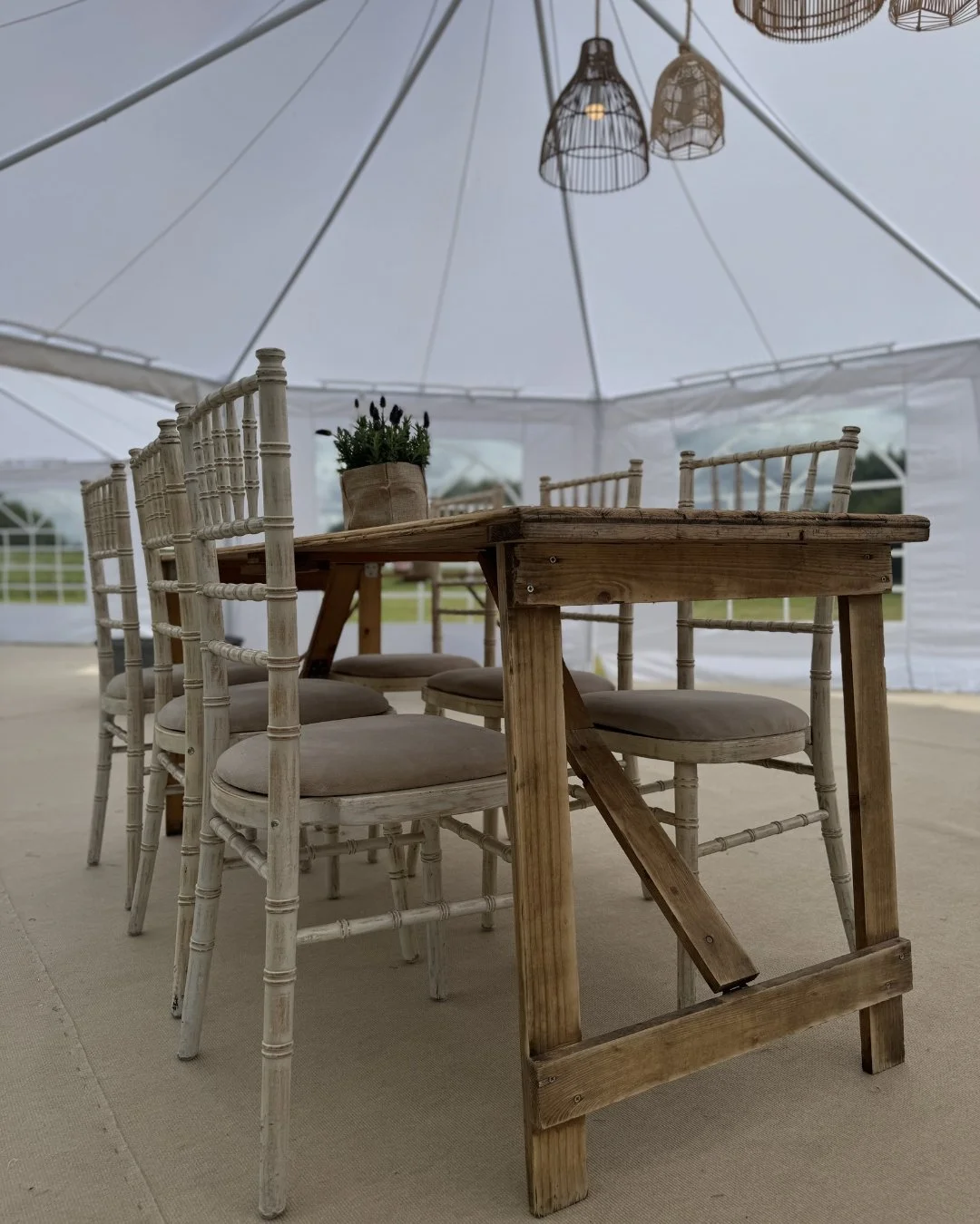 A picture of a rustic trestle table alongside chiavari limewash chairs in a traditional pole marquee