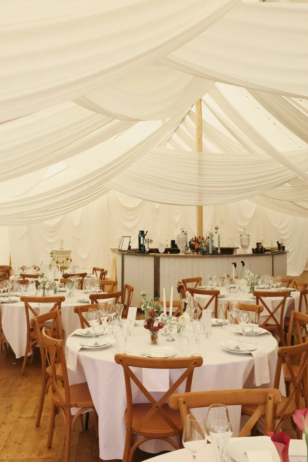 A traditional pole marquee with white drapes, round tables and crossback chairs decorating the wedding venue.