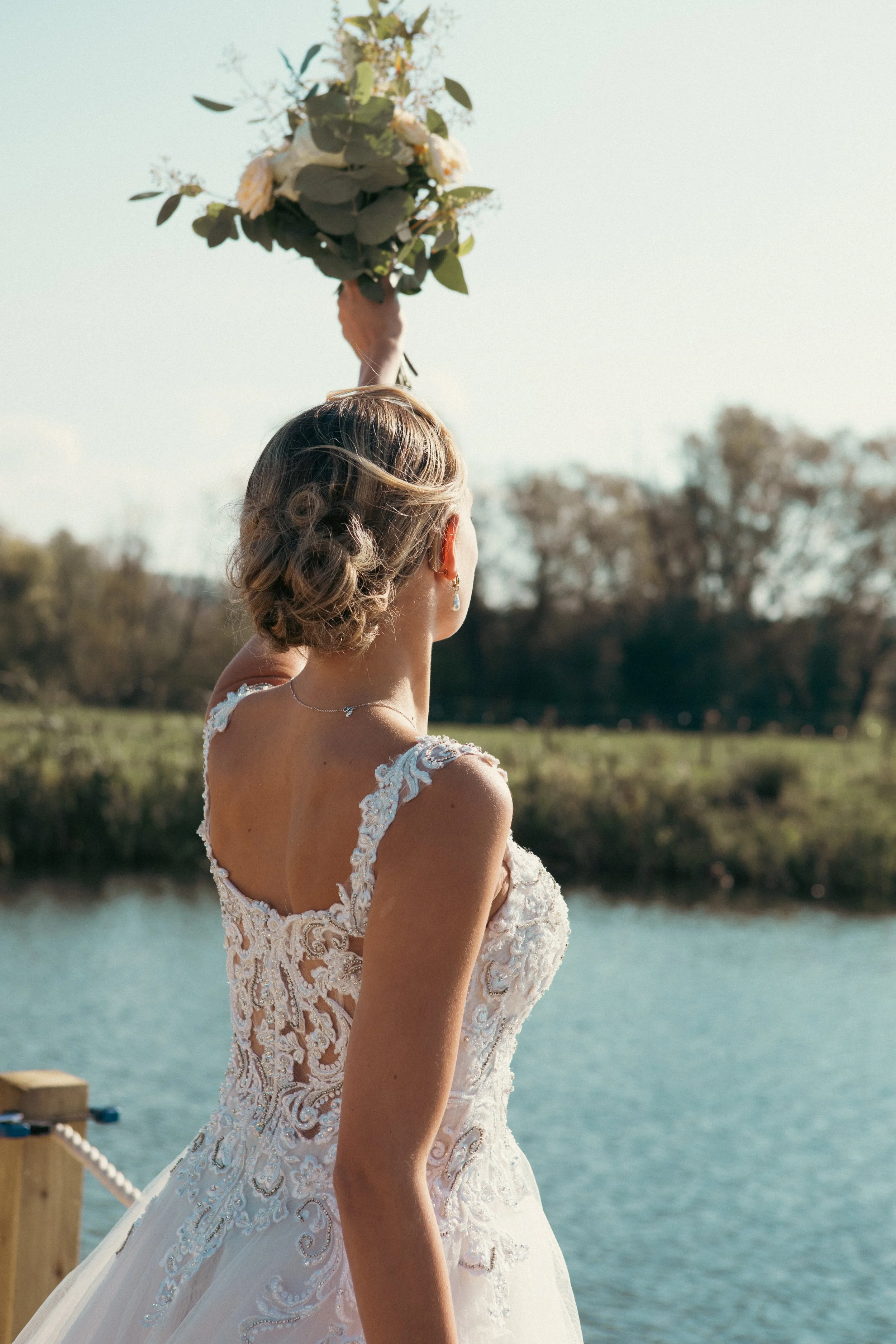 A picture of a bride wearing a wedding dress with her back to the camera holding a bouquet of flowers in the air at a lake.