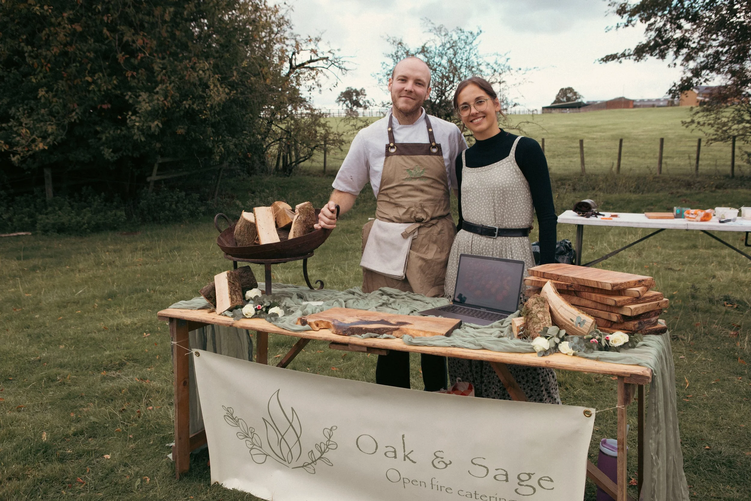 Outdoor open-fire catering setup with two people in aprons, firewood, wooden serving boards, and a laptop in a countryside field.