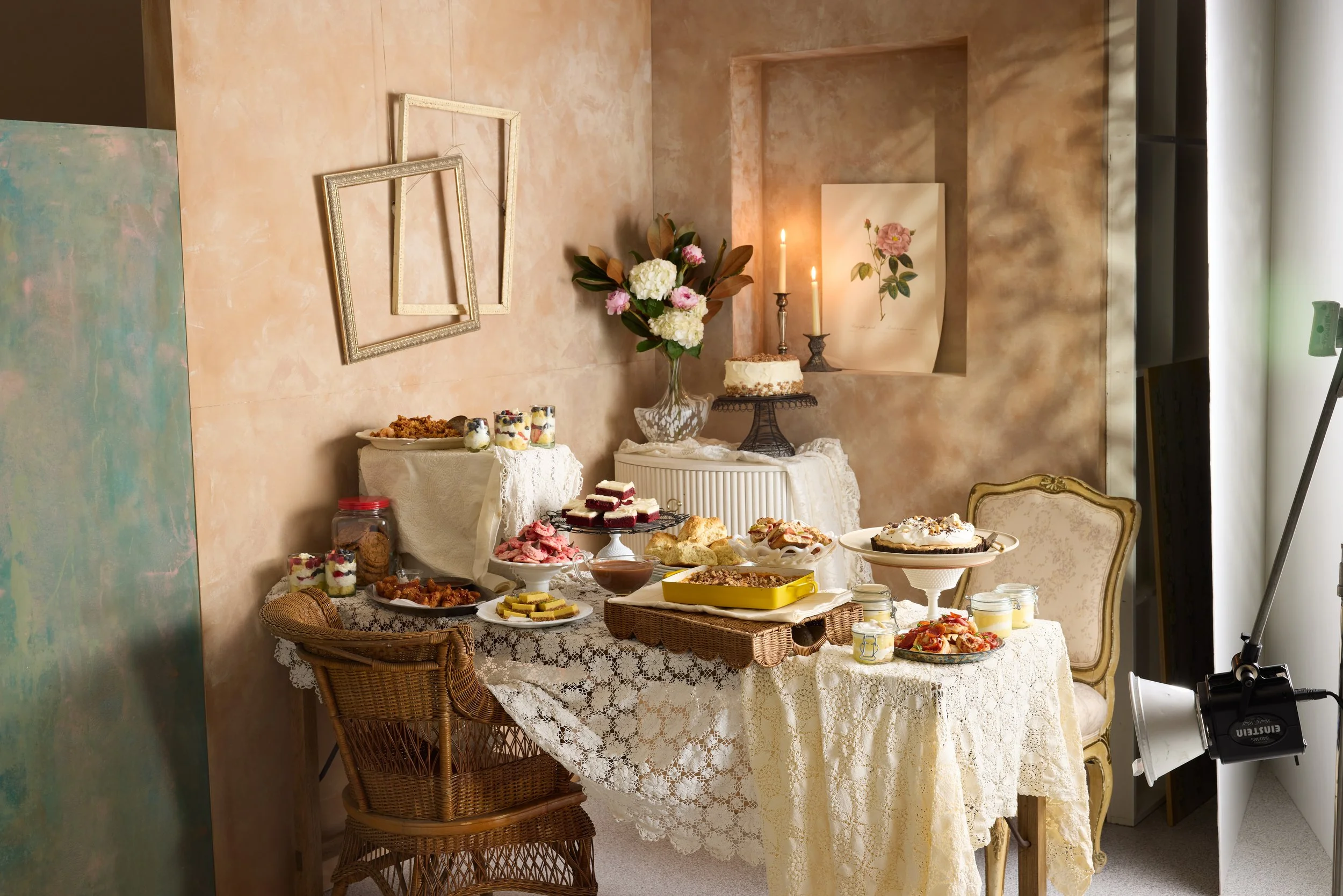 A decorated dining table with assorted desserts, surrounded by vintage chairs, set against a warm-toned wall with empty picture frames, flowers, and lit candles.