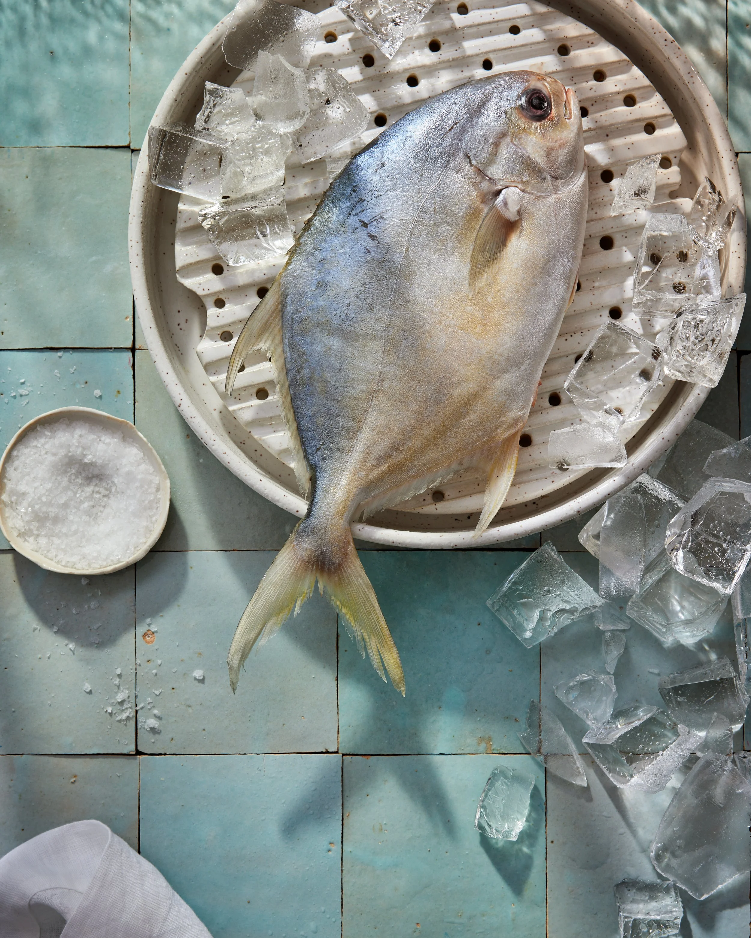 Fresh fish on a bed of ice in a wire basket, with a small bowl of salt nearby and ice scattered on a tiled surface.