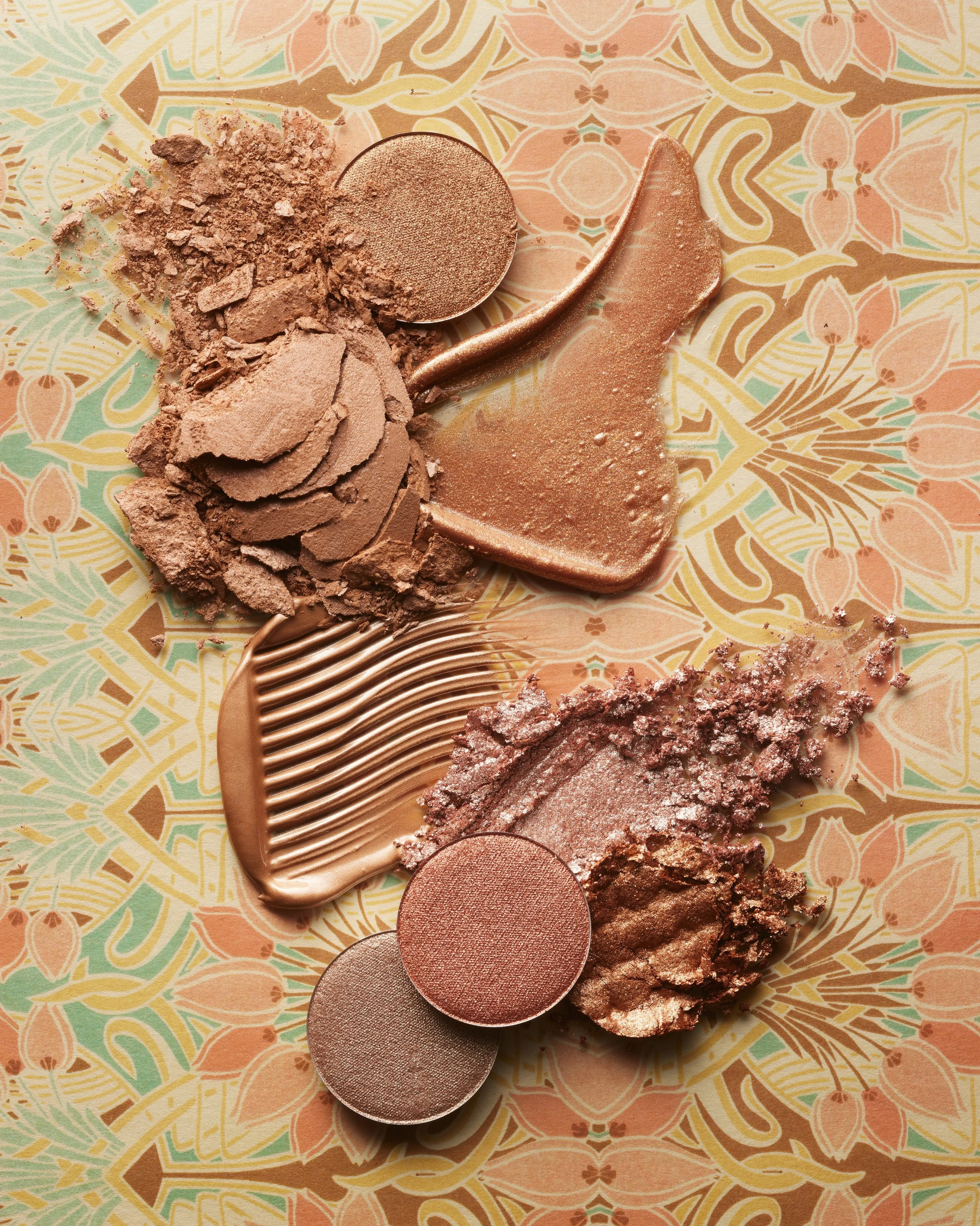 Various shades of shattered and whole makeup eyeshadows in bronze, pink, and nude tones on a patterned surface.