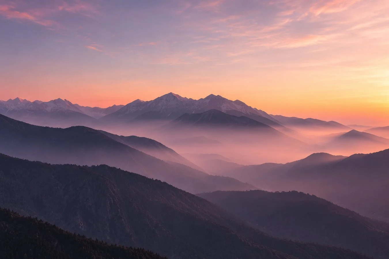 Sunset over a mountain range with layers of mist and snow-capped peaks in the distance.