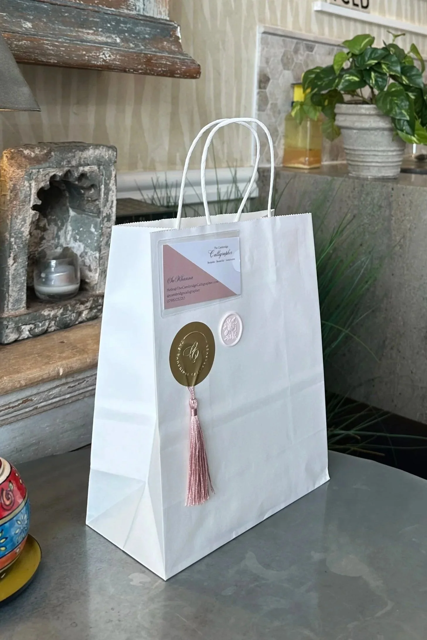 White paper shopping bag with business card and decorative seal attached, placed on a gray countertop in a kitchen or dining area.
